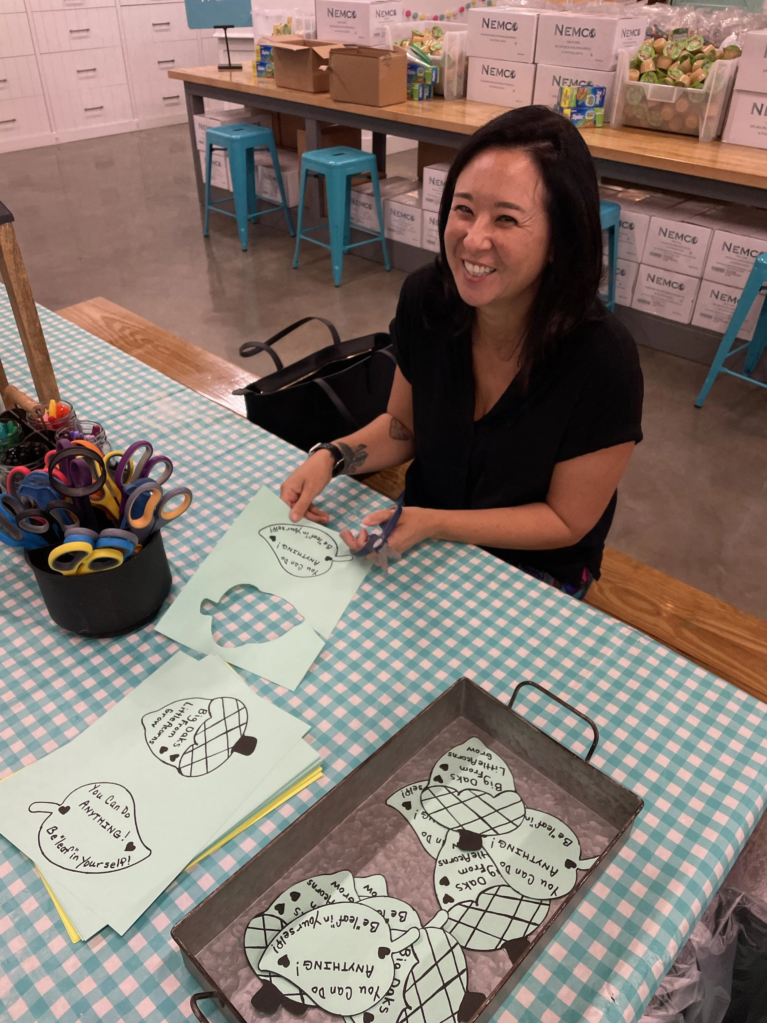 A woman sitting at a table with crafting supplies, including scissors, paper, and cutouts with motivational messages, in a room with storage boxes and high chairs in the background.
