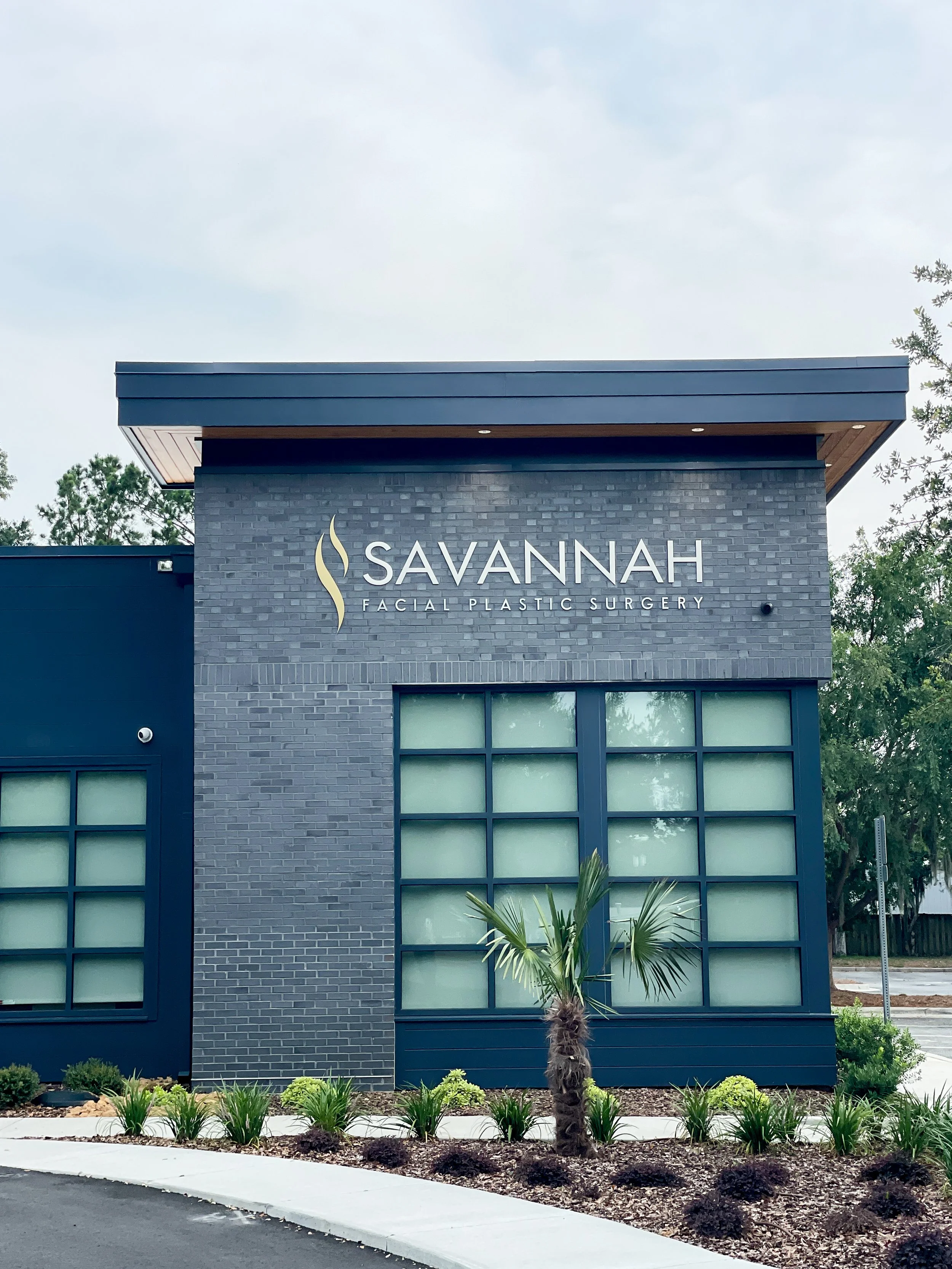 Exterior of Savannah Facial Plastic Surgery clinic, modern building exterior with dark brick and large windows, small palm tree and shrubbery in front.