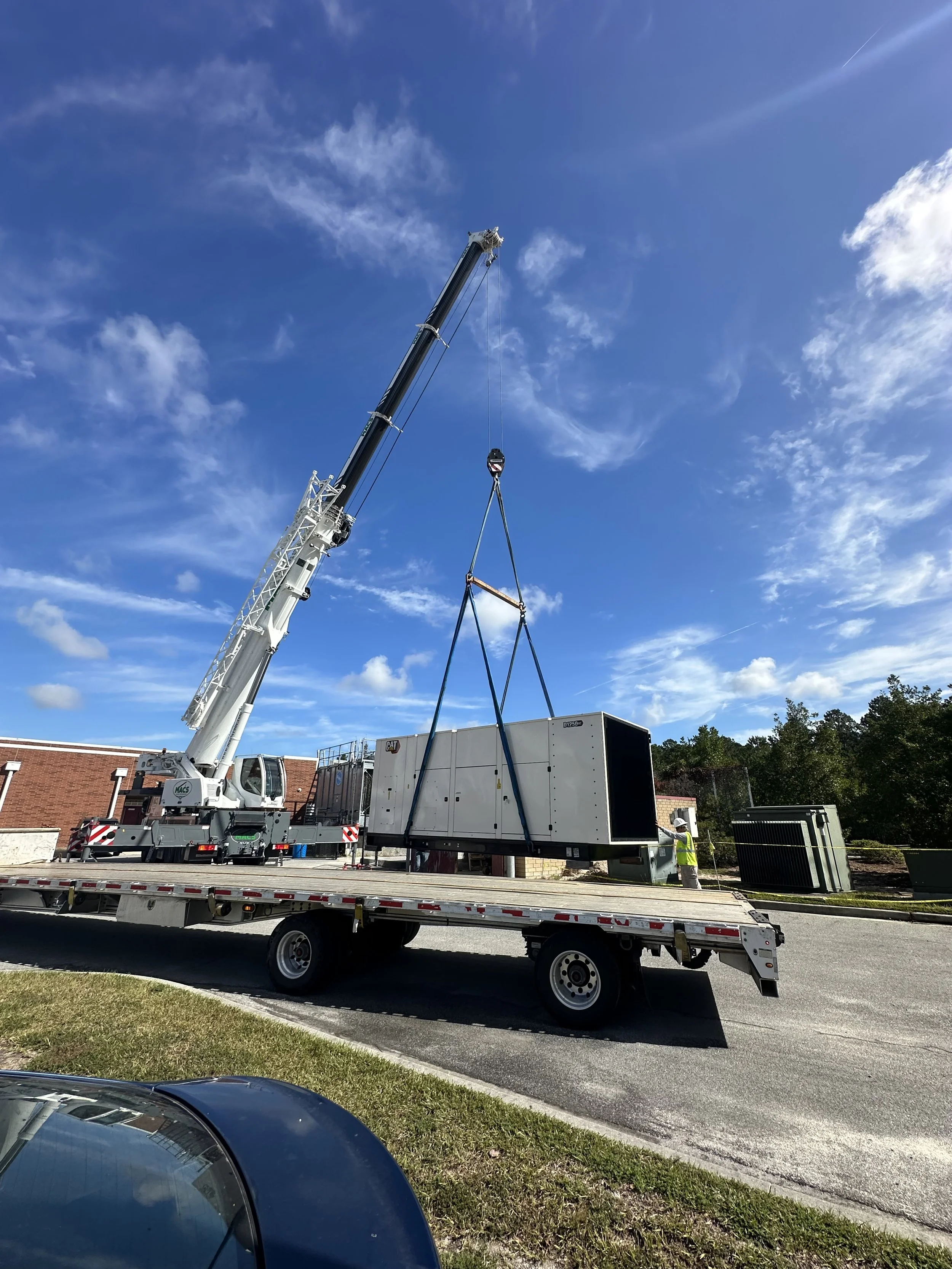 A crane on a flatbed truck lifting a large black and white industrial HVAC unit or similar equipment outdoors on a clear, sunny day.