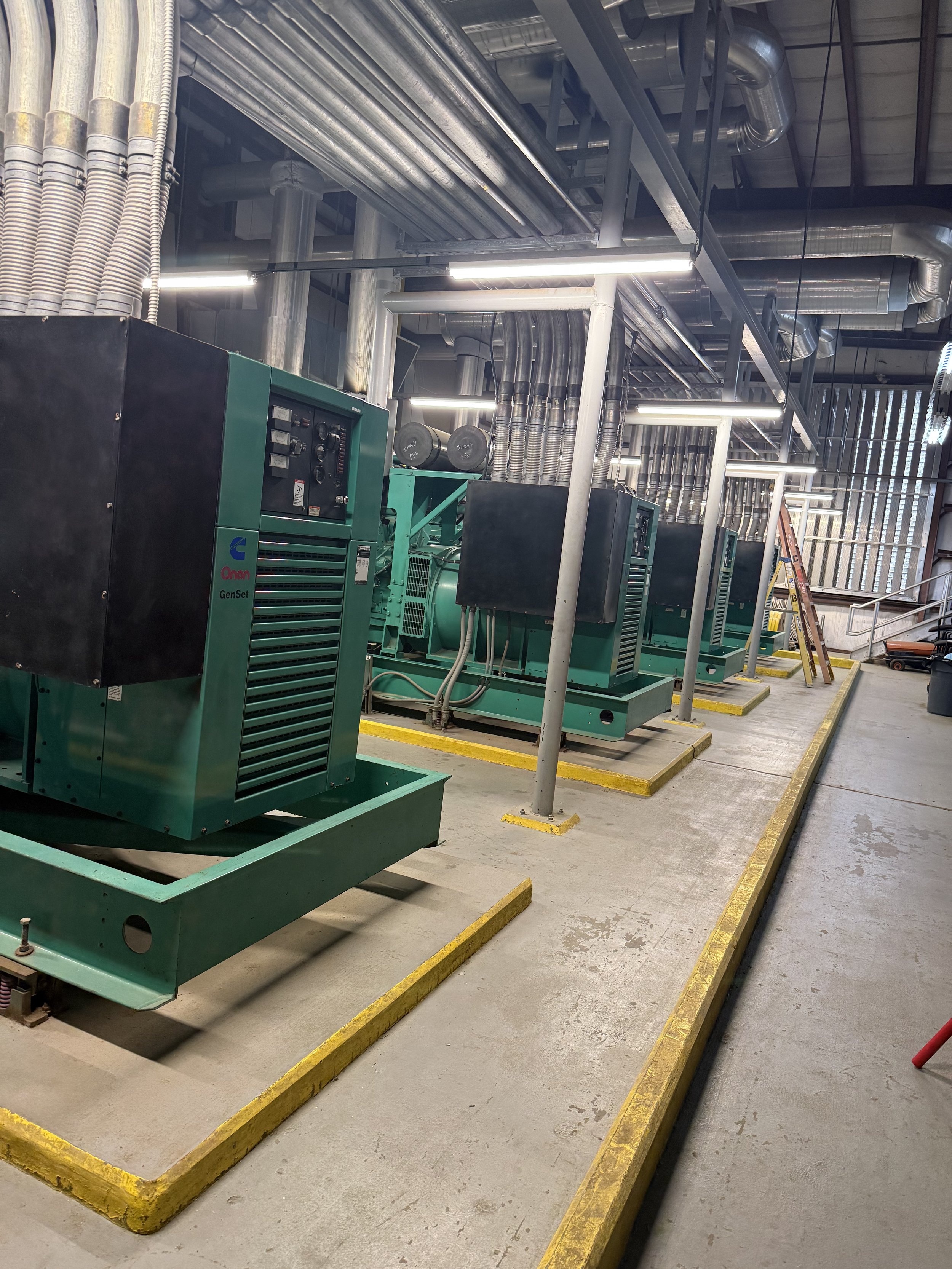 Industrial piping and generators in a power plant or data center, with yellow safety barriers on the concrete floor.