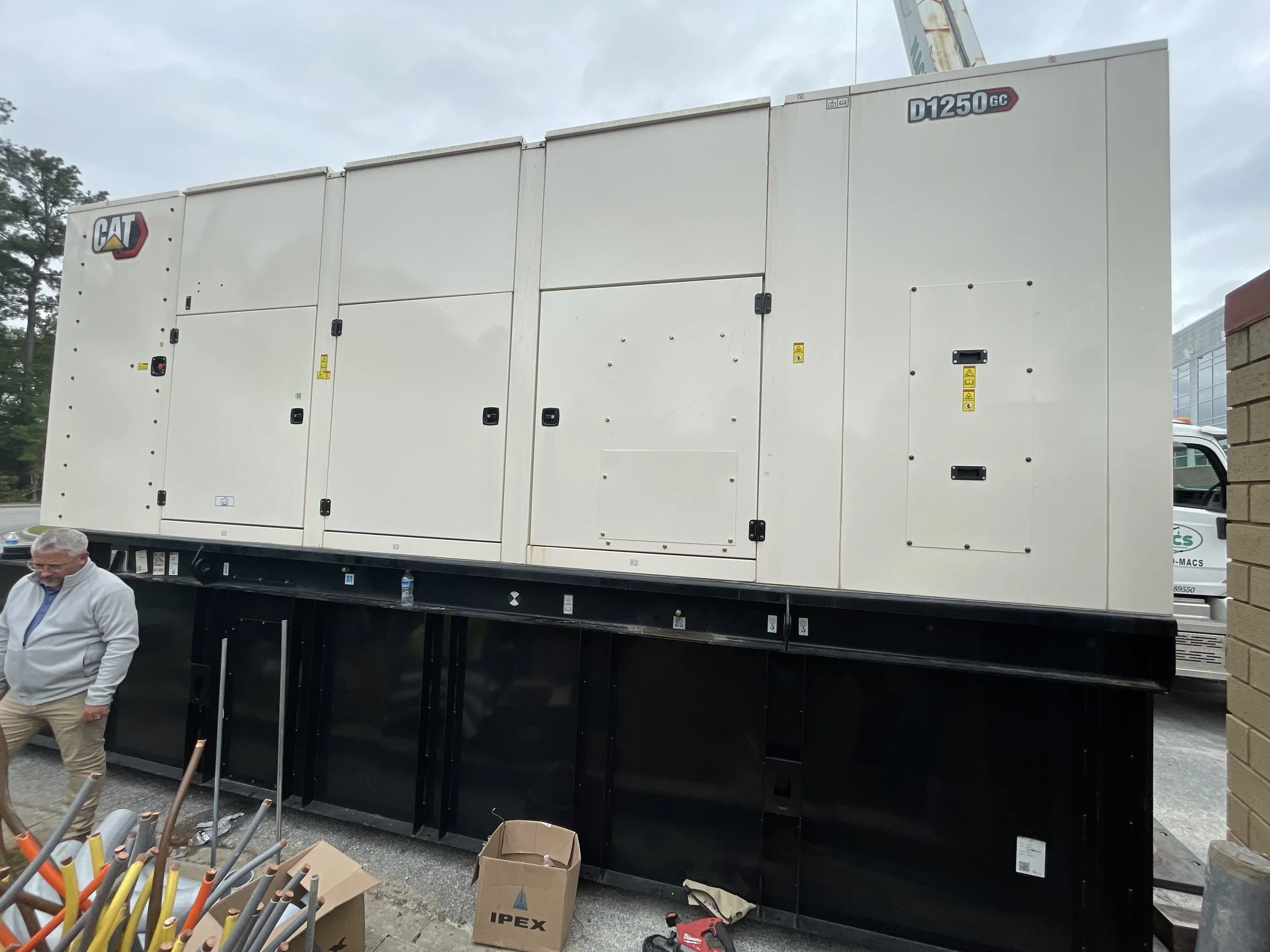 A large white industrial generator with the CAT logo on it, mounted on a black metal platform, next to a brick wall and a man standing nearby.