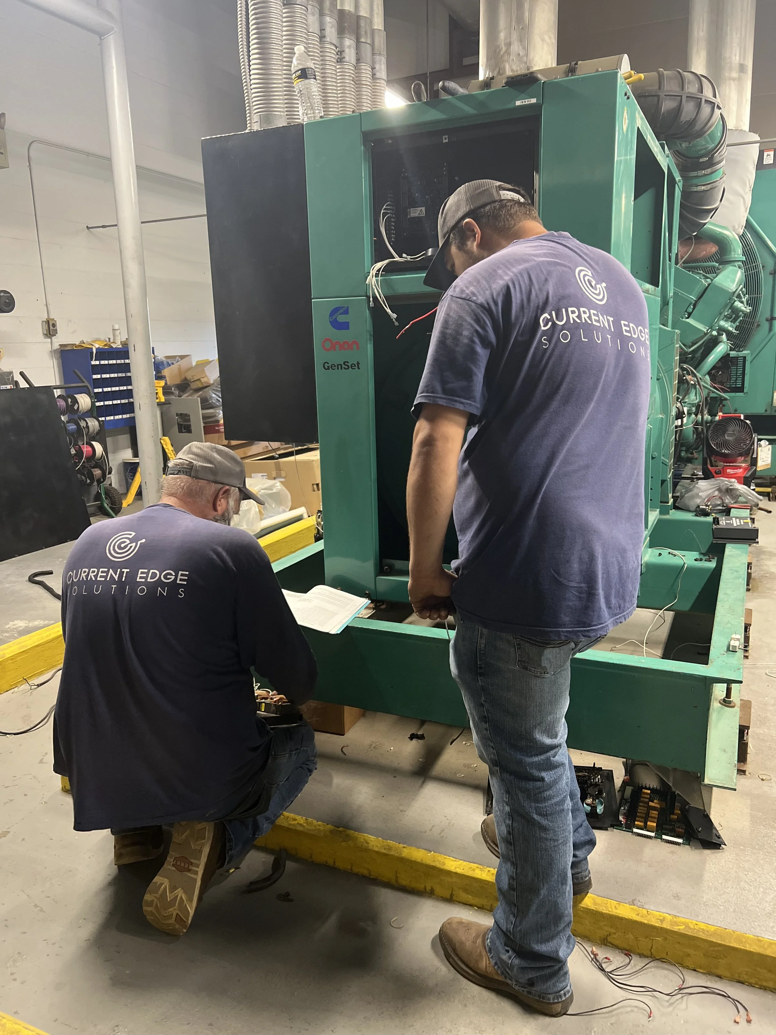 Two workers in a factory or workshop working on a large green machine. One worker is kneeling and looking at a manual, while the other is standing and working on the machine. Both are wearing navy blue t-shirts with "Current Edge Solutions" printed o