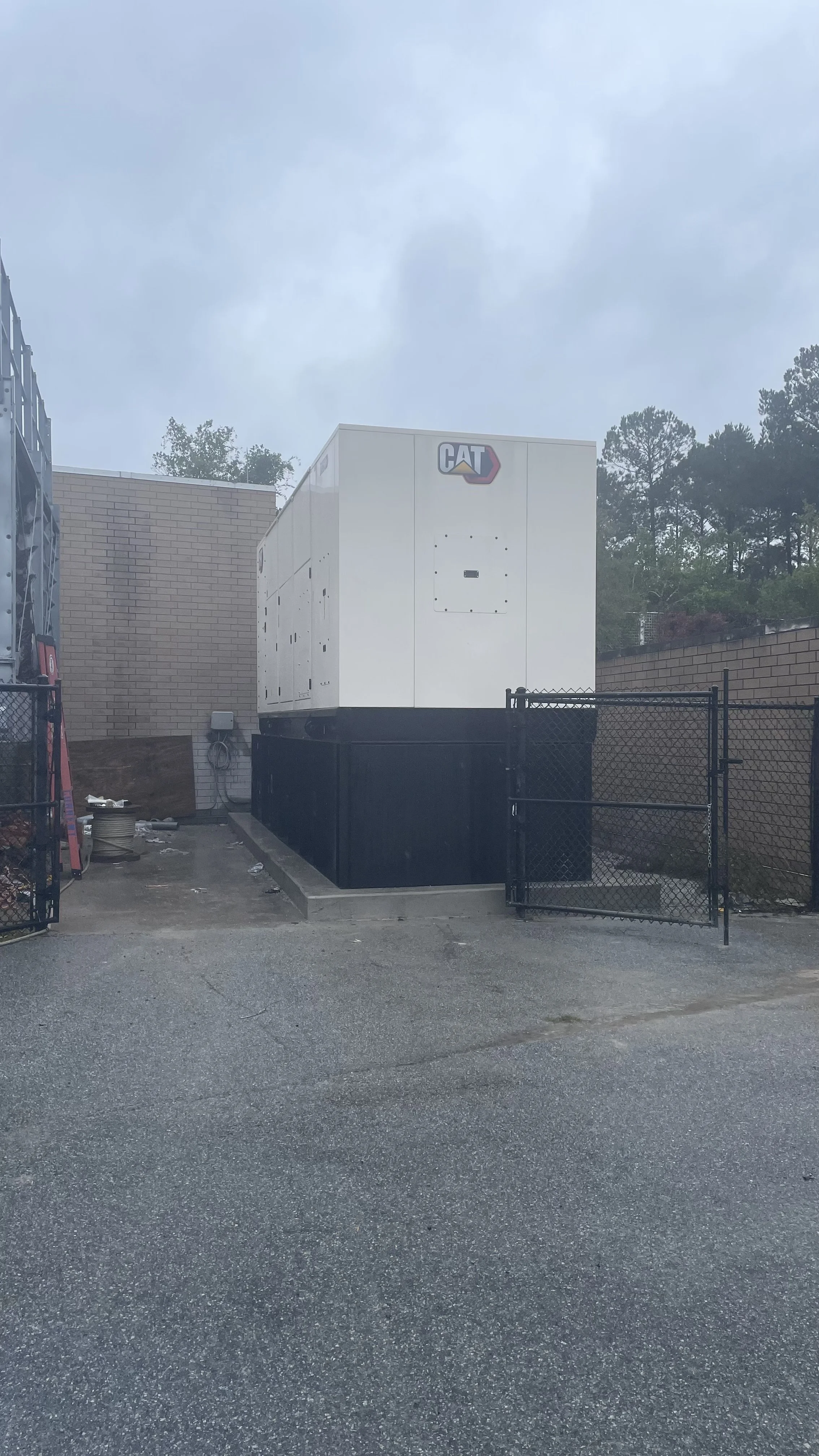 Large white Caterpillar generator outside, surrounded by a black fence, against a brick wall with cloudy sky overhead.