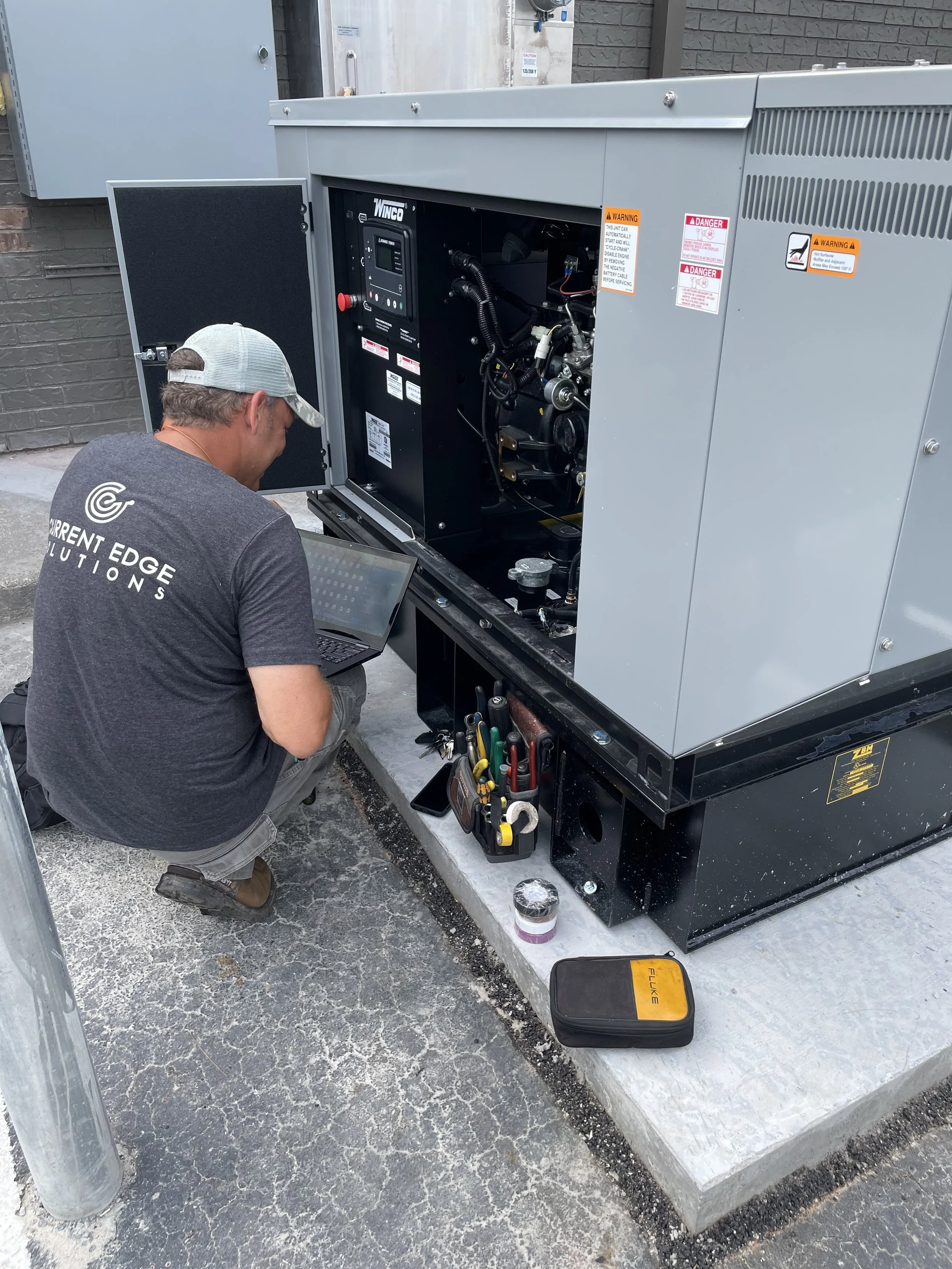 Technician working on a large industrial generator, inspecting the control panel and internal components outside on a concrete pad.
