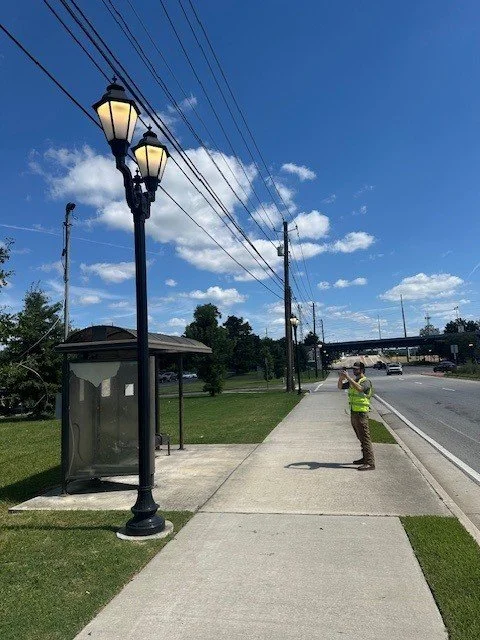 A person in a yellow safety vest standing on a sidewalk, taking a photo or video with a smartphone. The scene includes a street with a bus stop shelter, a decorative lamppost, utility poles, and power lines under a partly cloudy sky.