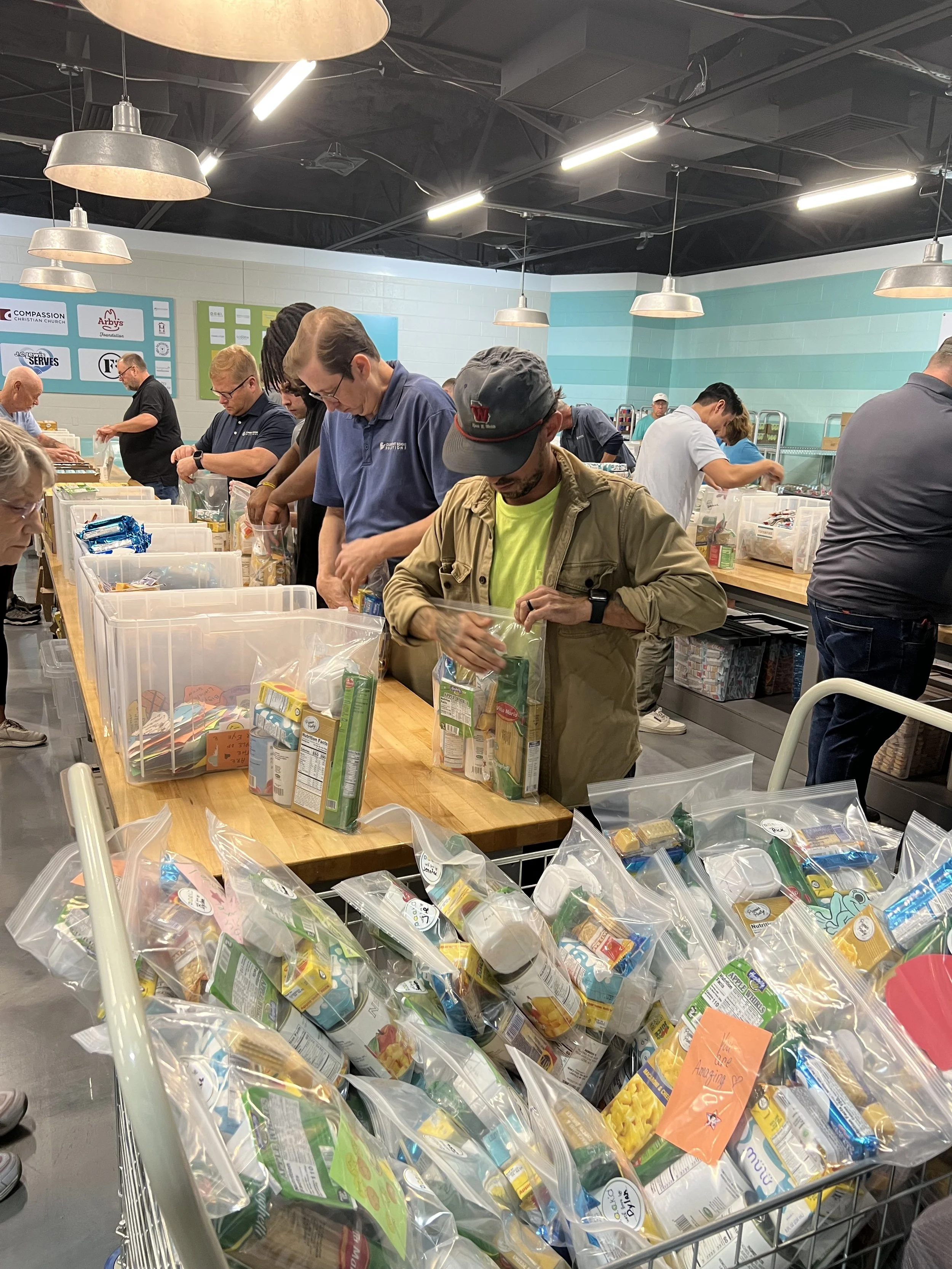 People packing food into plastic bags at a community service event in a well-lit room.