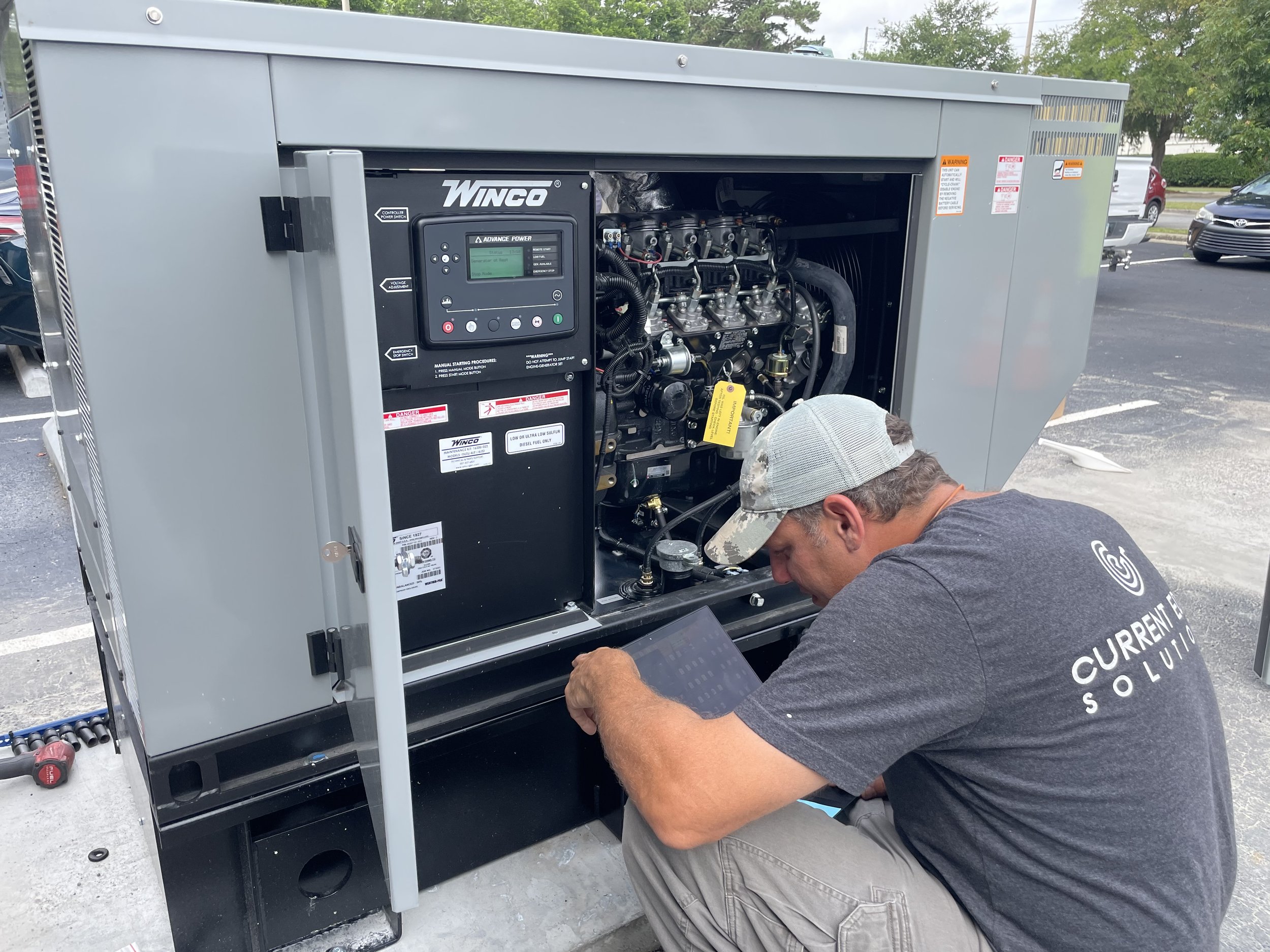 A technician is inspecting or working on a large industrial generator outdoors. The technician is using a tablet or electronic device while kneeling in front of the open metal enclosure of the generator, which reveals its internal components.
