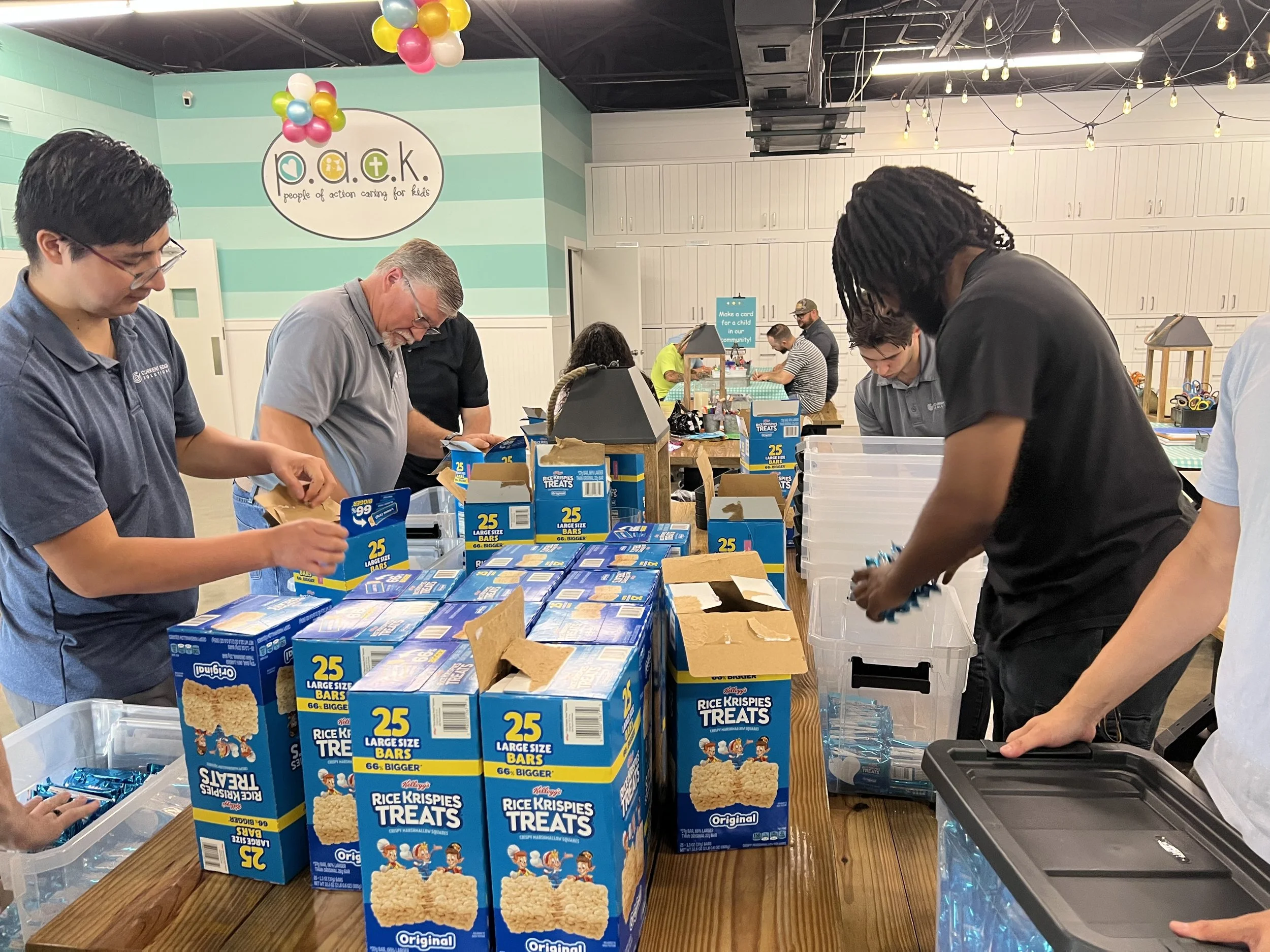 People packing boxes of Rice Krispies Treats in a room decorated with balloons and a sign reading "People of Action Caring for Kids."