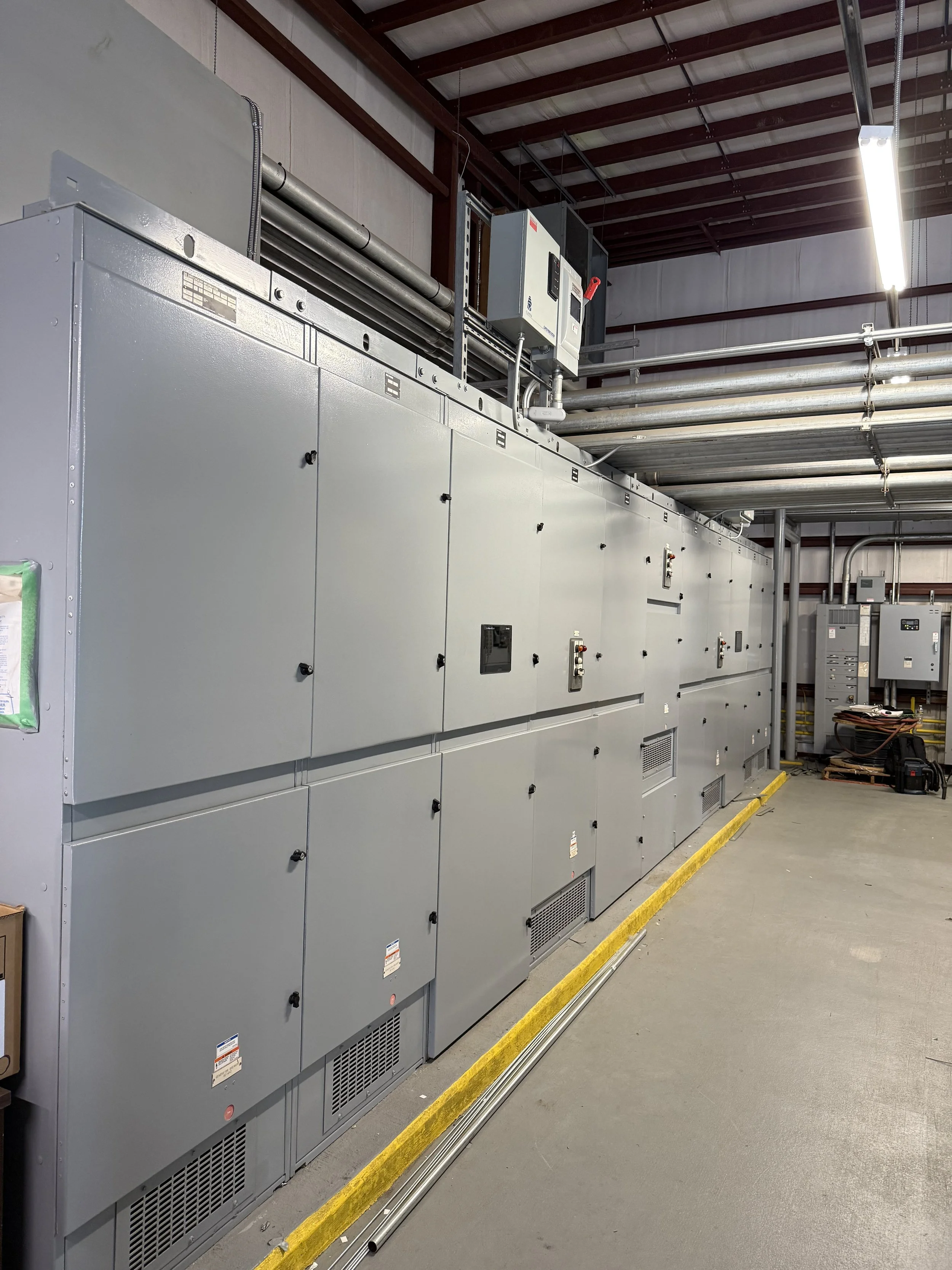 Electrical control panel in an industrial setting with gray metal cabinets, pipes, and wiring.