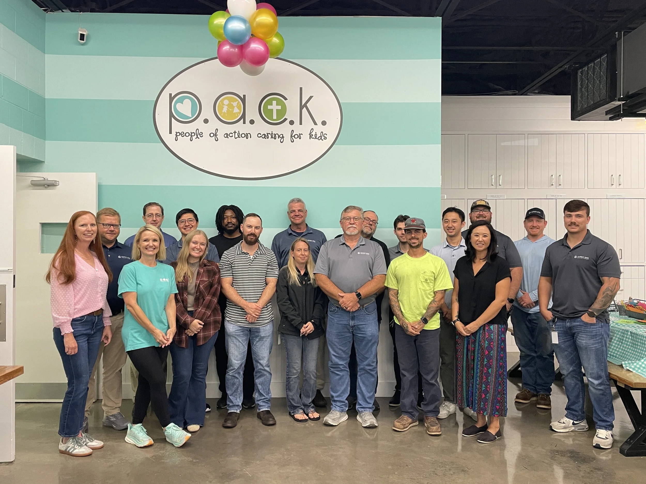 Group photo of people standing indoors in front of a teal and white striped wall with a logo that reads 'p.a.c.k. People of Action Caring for Kids' and a cluster of colorful balloons above the logo.