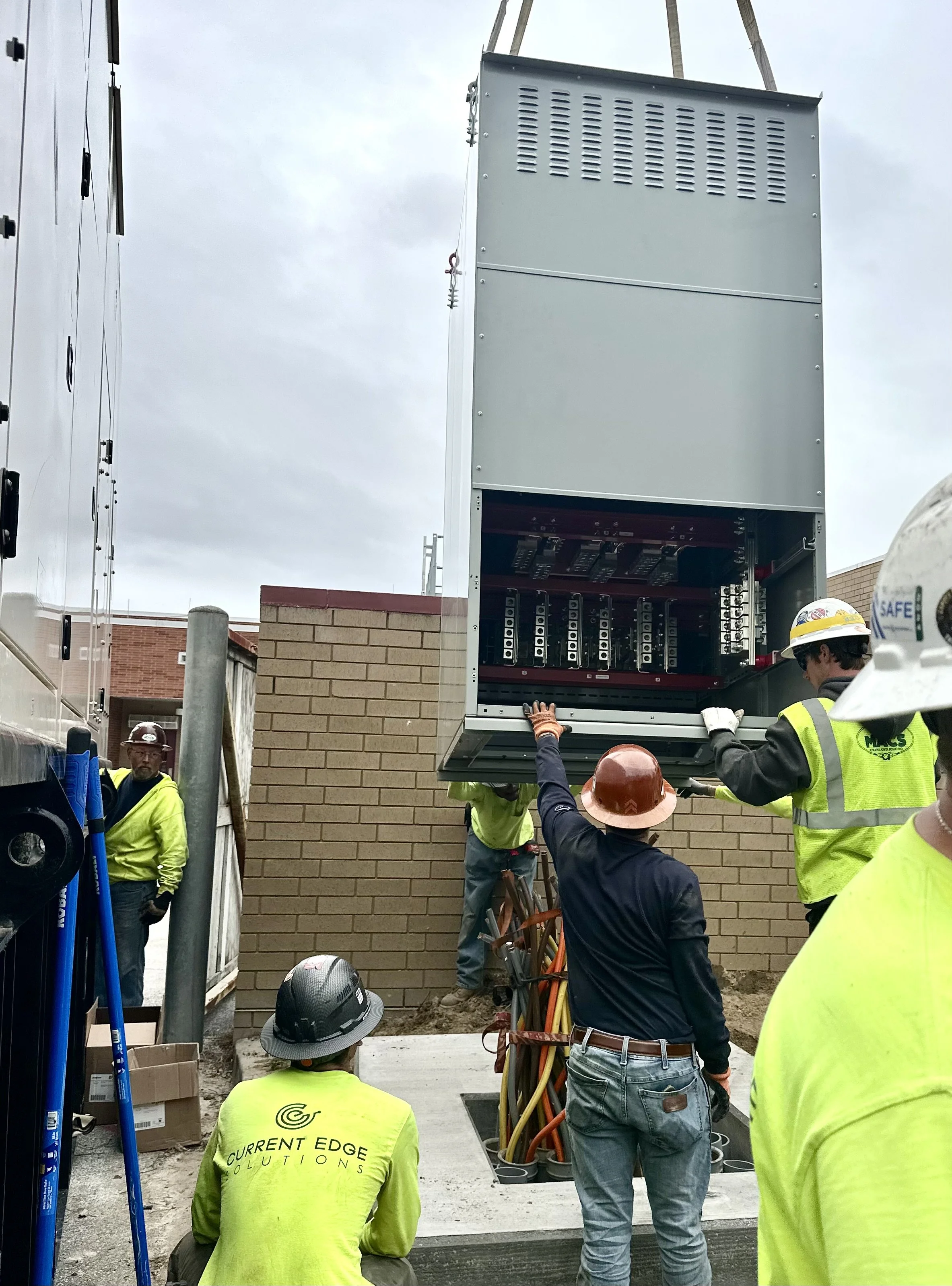 Construction workers install electrical equipment in a large metal electrical box outside a building, with some workers wearing safety gear like helmets and reflective vests.