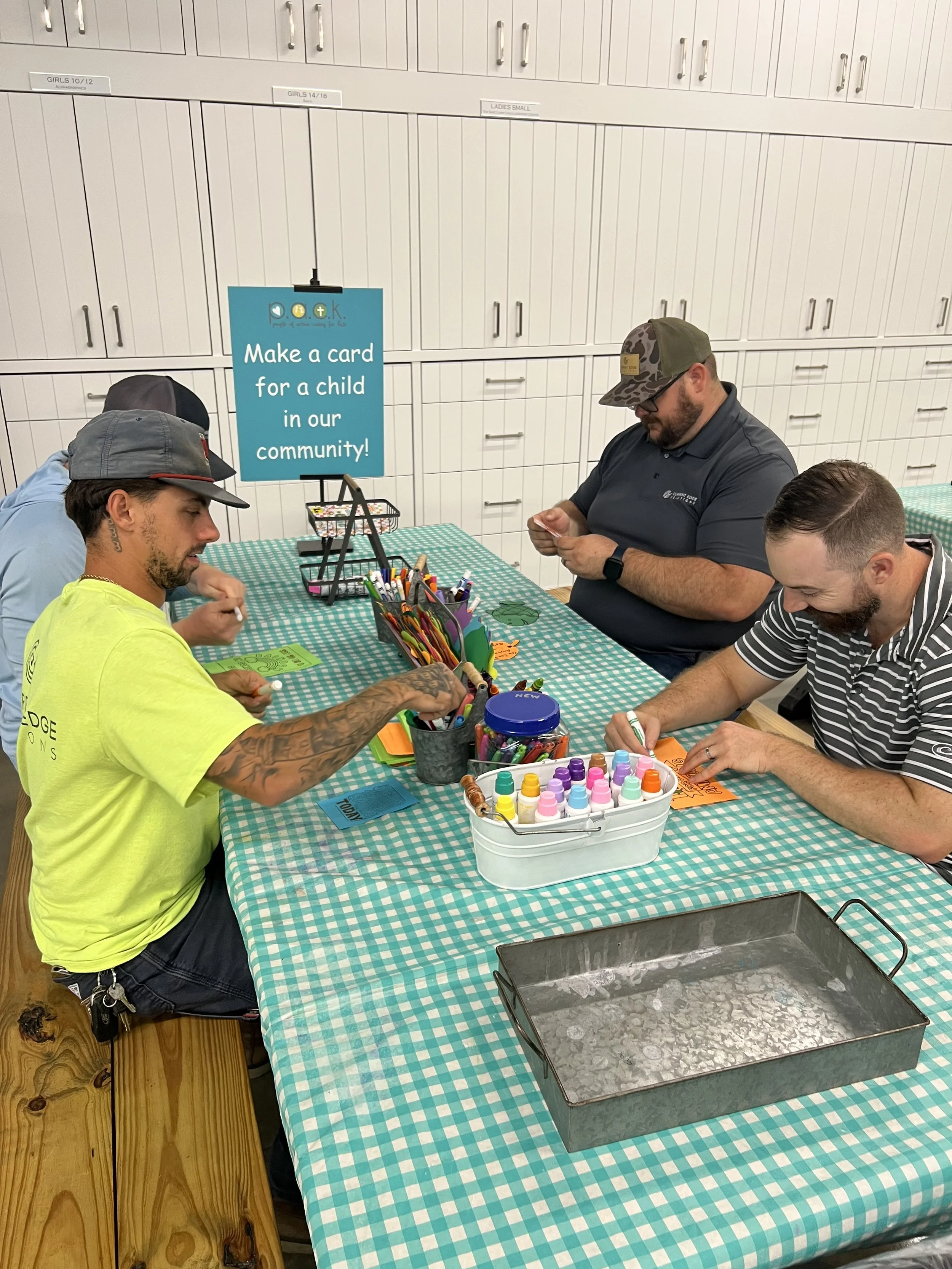 Four men sitting at a table making cards for children in the community. The table has a green and white checkered tablecloth, and supplies like markers, paint, and paper are on it. A sign on a stand reads, 'Make a card for a child in our community!'