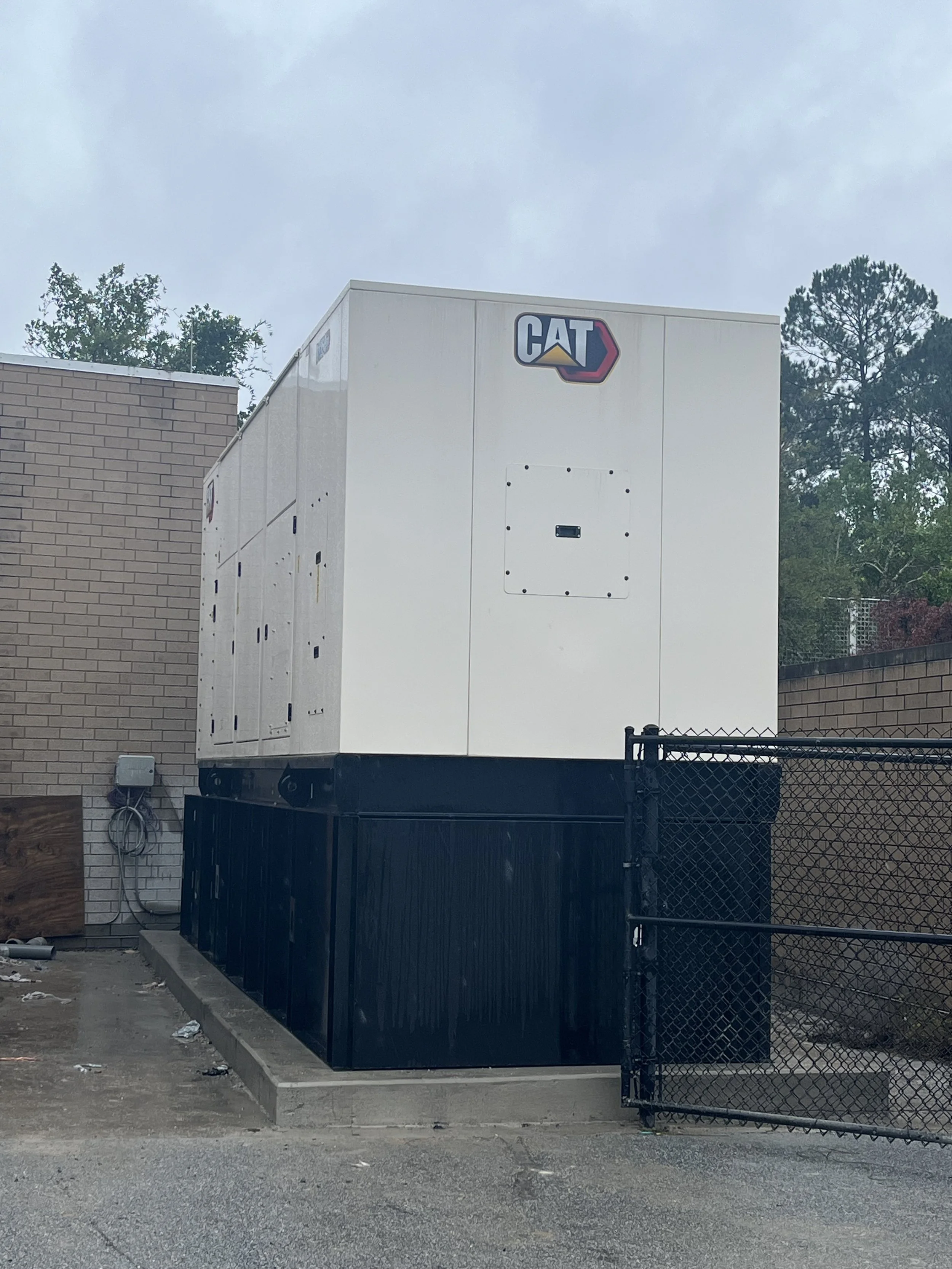 Large white generator with CAT logo on top, situated next to a brick wall and black fence, in an outdoor industrial area.
