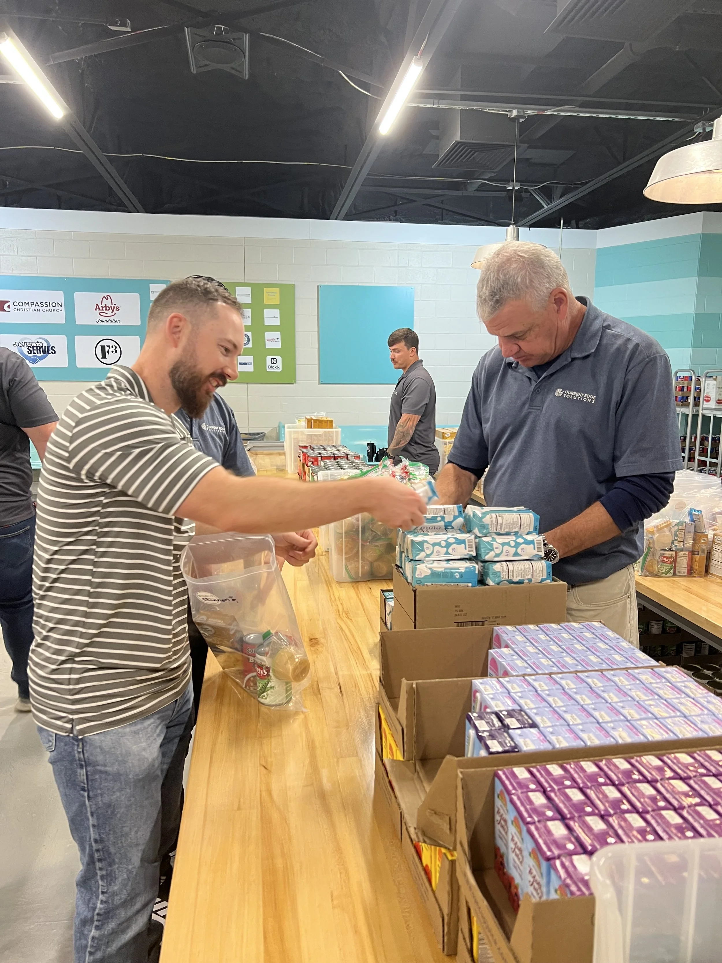 Two men packing food items into bags at a community service event, with one man smiling and the other focused on organizing products on a wooden table.