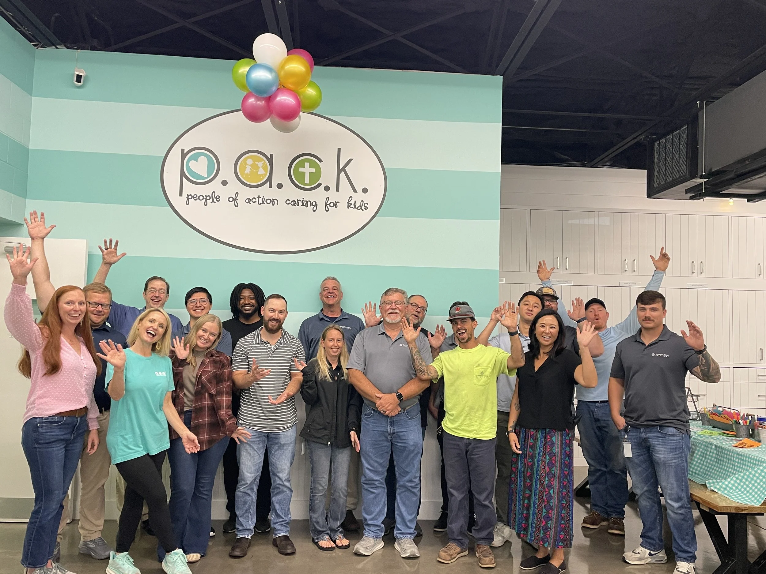 Group of smiling people posing together with arms raised inside a room with a mint green wall, a sign that says p.a.c.k. (people of action caring for kids), and balloons hanging from the ceiling.