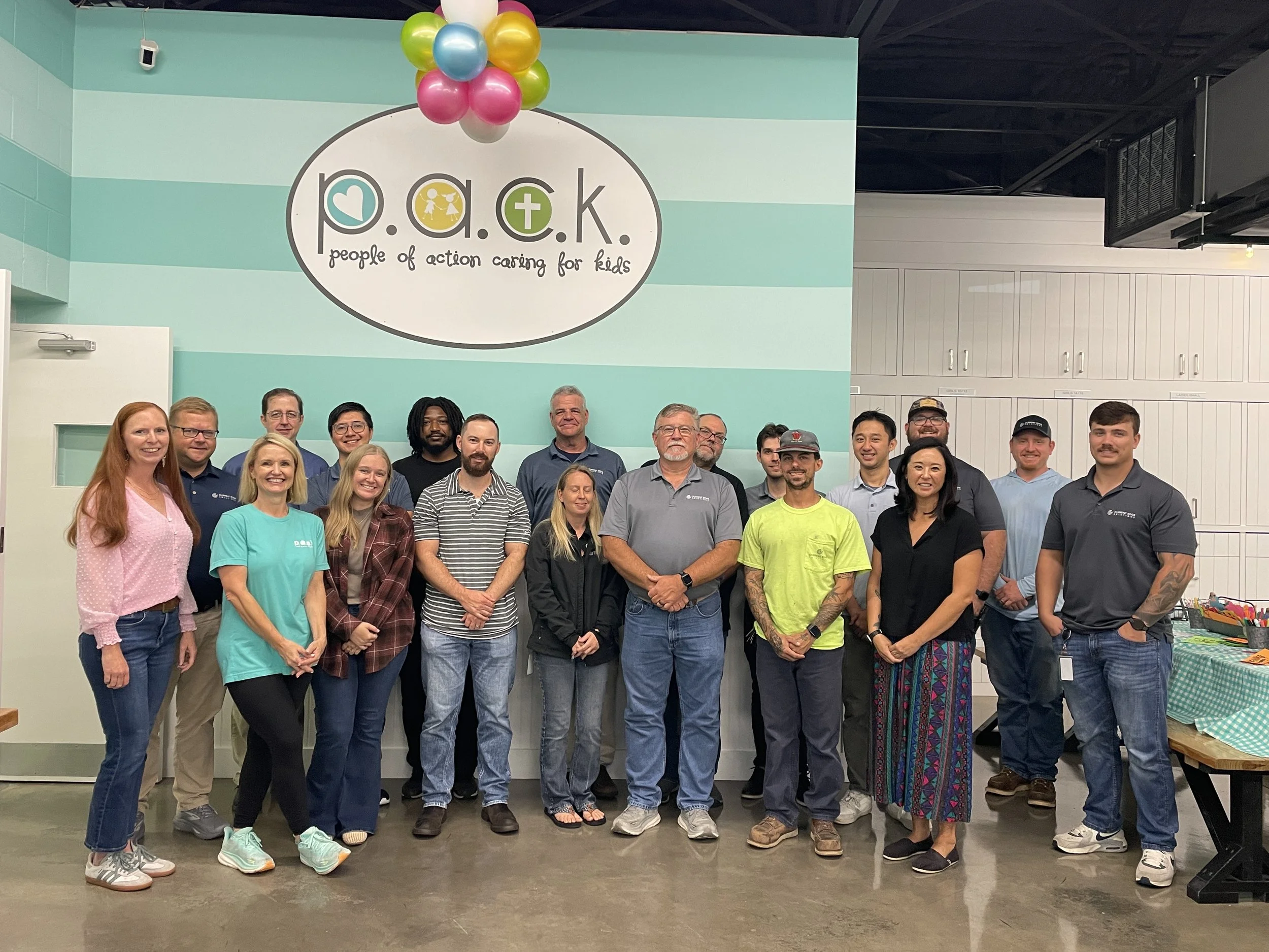 Group of diverse adults standing inside a room with a turquoise wall, smiling for a photo, in front of a sign that says 'P.O.A.C. people of action caring for kids,' with colorful balloons above it.