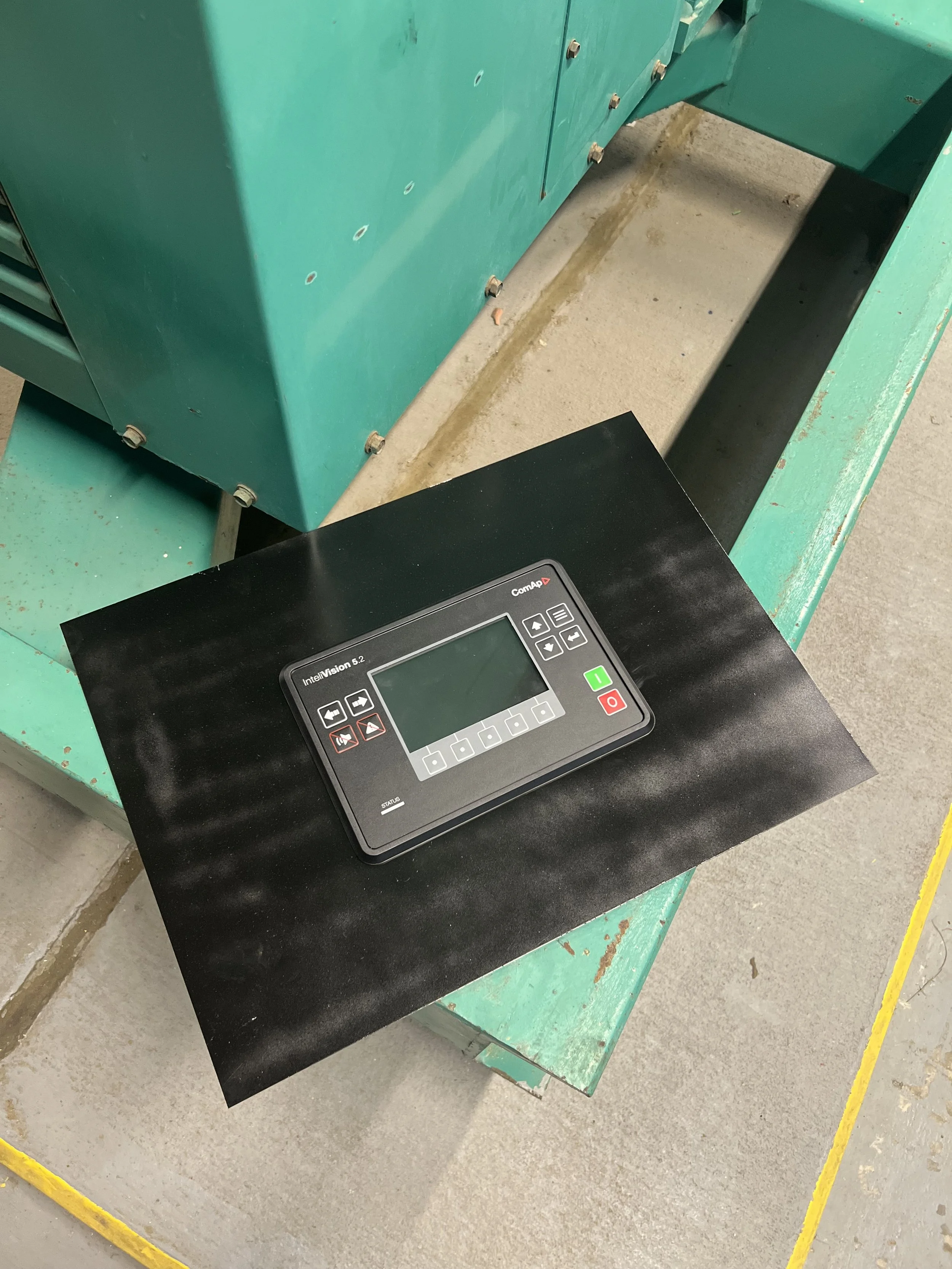 A black control panel with a digital screen and buttons resting on a black surface. The background shows green equipment and a concrete floor.