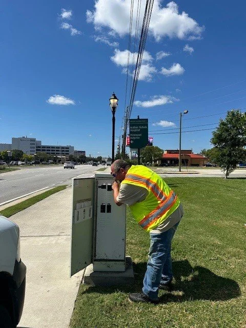 A man in a yellow safety vest leaning over and looking into an outdoor electrical box next to a sidewalk on a sunny day.