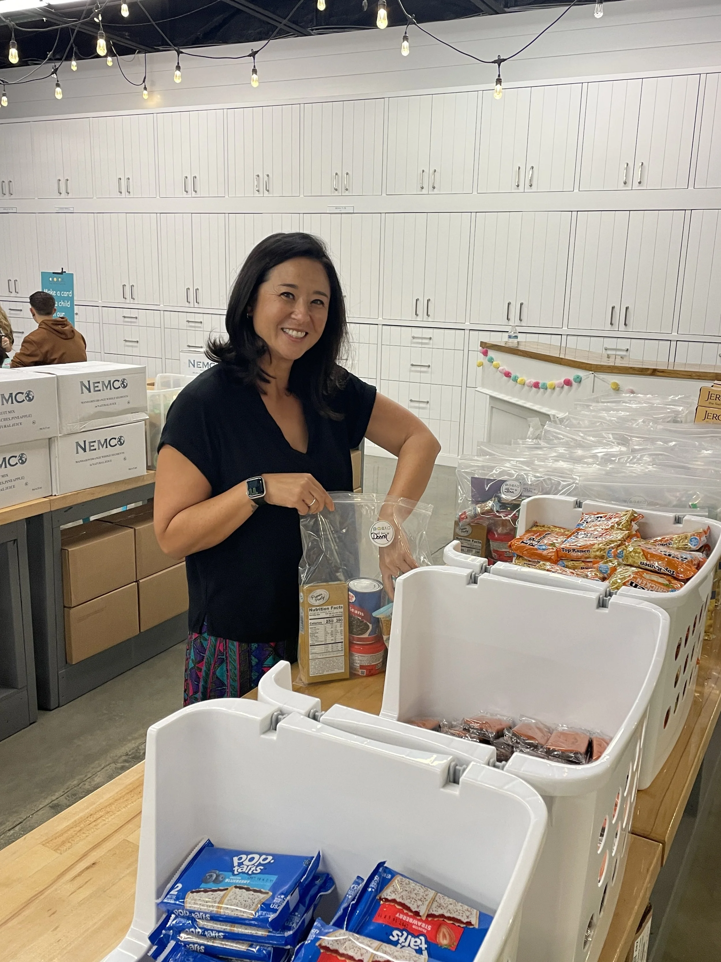 Woman smiling at camera while standing behind table with food packages, inside a room with white cabinets and string lights overhead.