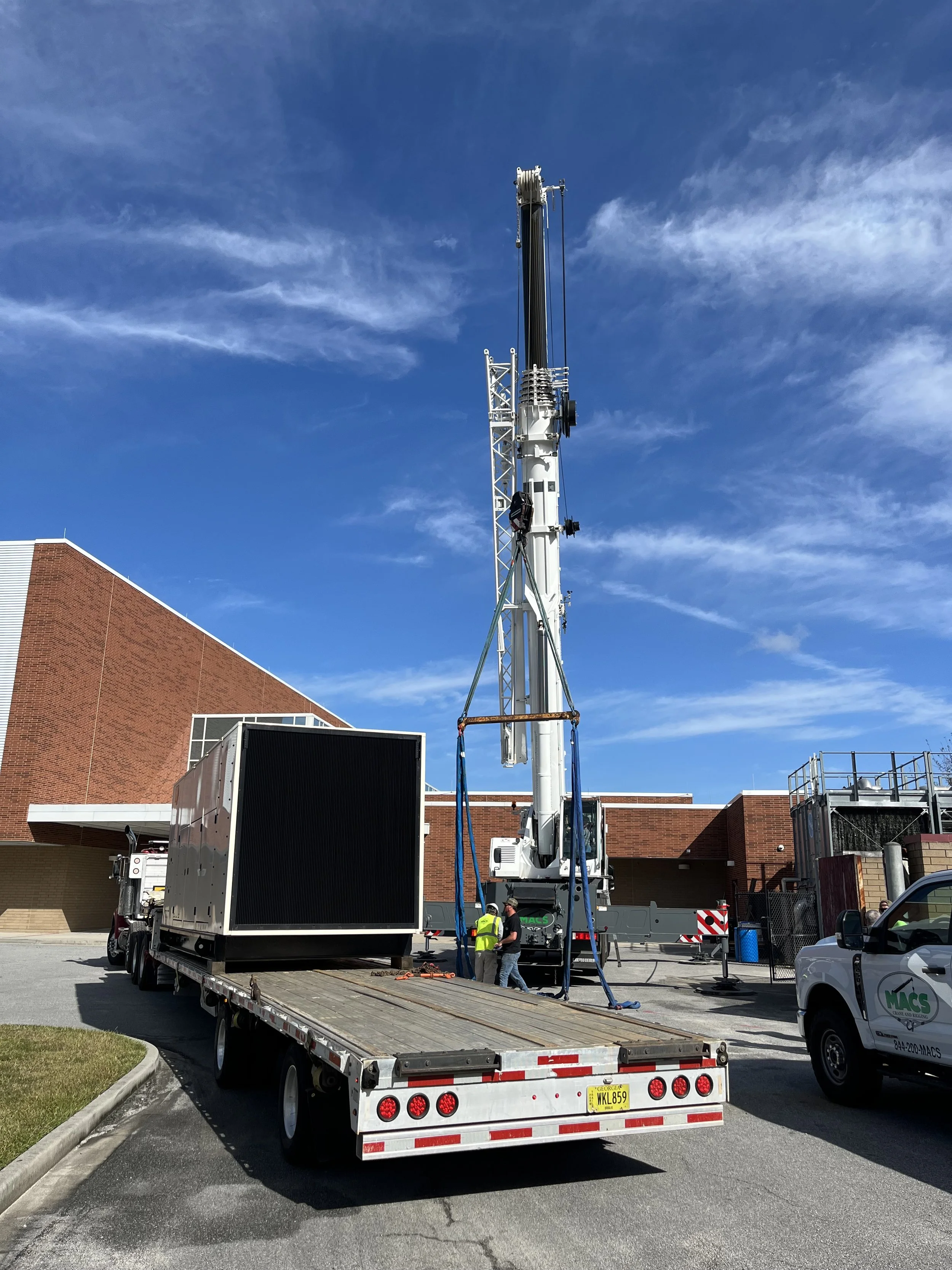 A crane lifting a large black rectangular object onto a flatbed truck, with workers standing nearby, in an outdoor area with brick buildings and a blue sky with some clouds.