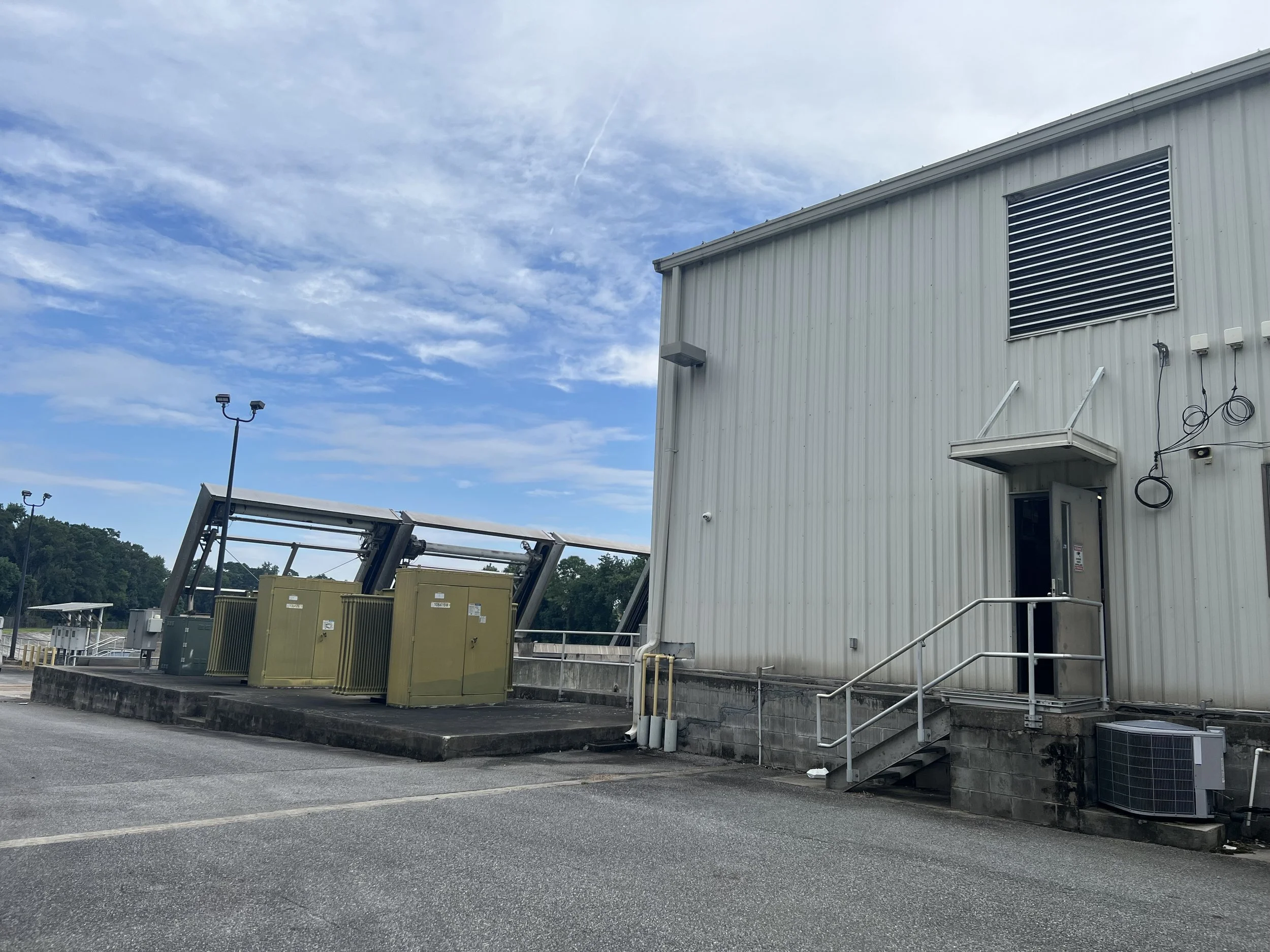 Exterior of a utility building with electrical boxes, transformers, and outdoor air conditioning unit, under a partly cloudy sky.