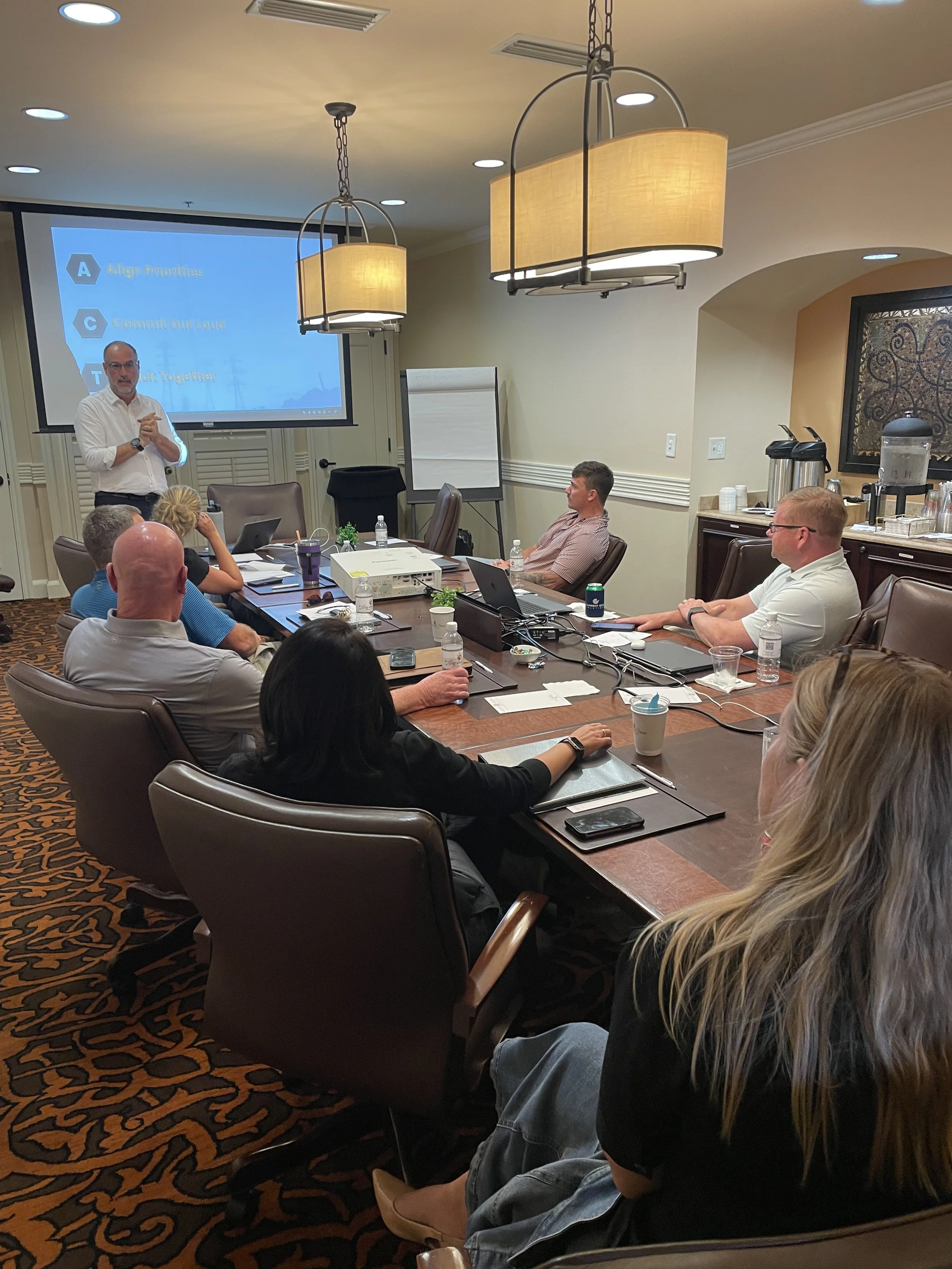 A business meeting or training session taking place in a conference room with several people seated around a large table, listening to a man standing and speaking in front of a projected presentation screen.