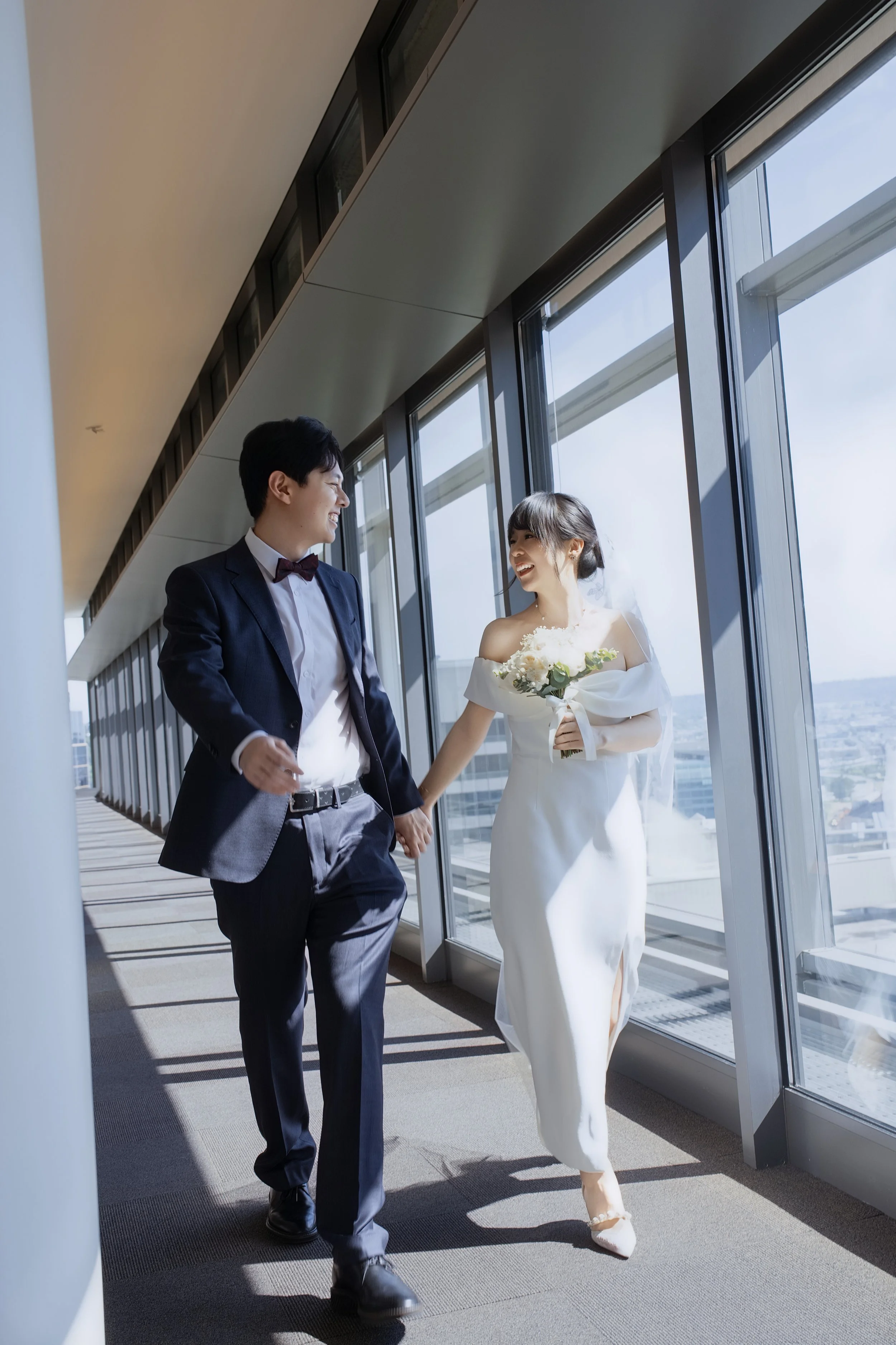 Smiling bride and groom holding hands and walking through a sunlit glass corridor