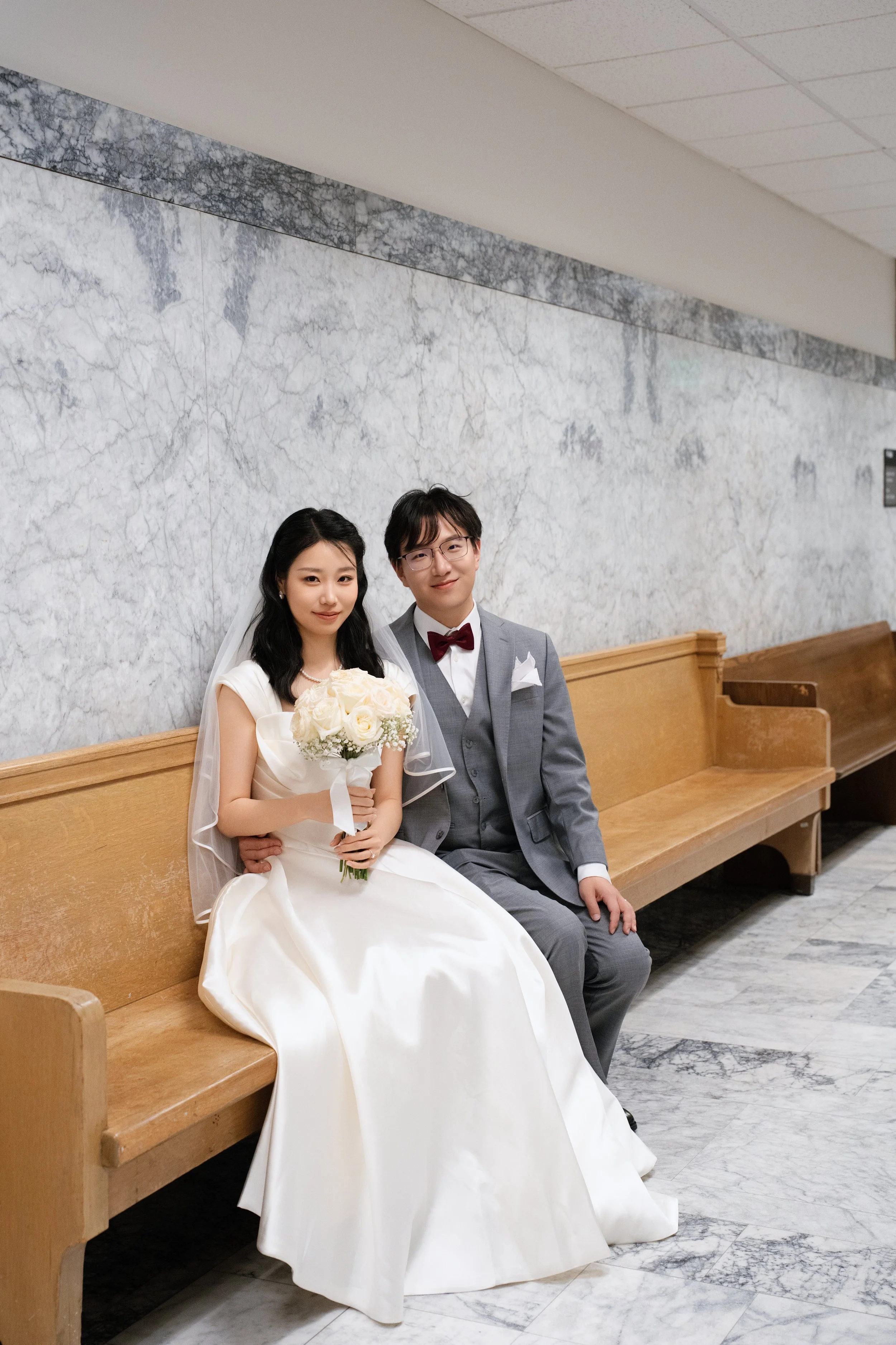 Newlywed couple seated in a church pew, with the bride holding a white rose bouquet and the groom wearing a gray suit and burgundy bow tie.