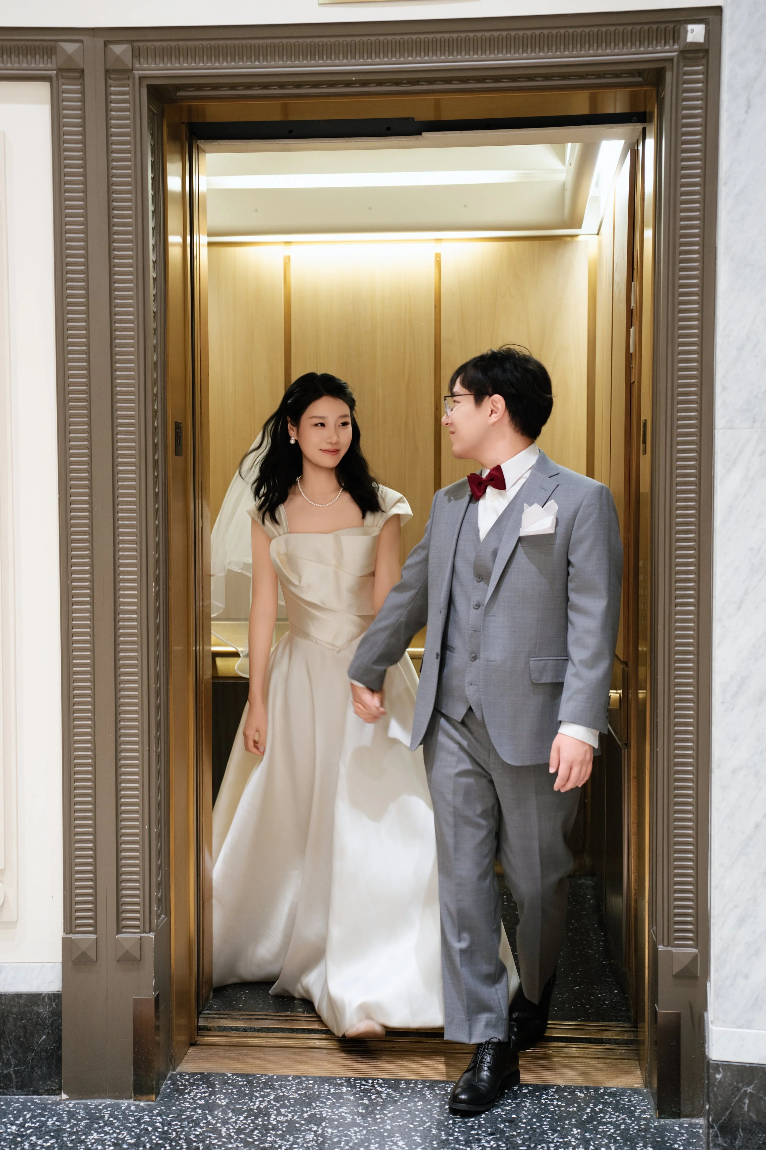 Newlywed couple posing in an ornate gold elevator, with the bride in a white wedding dress and the groom in a gray suit with a red bow tie.