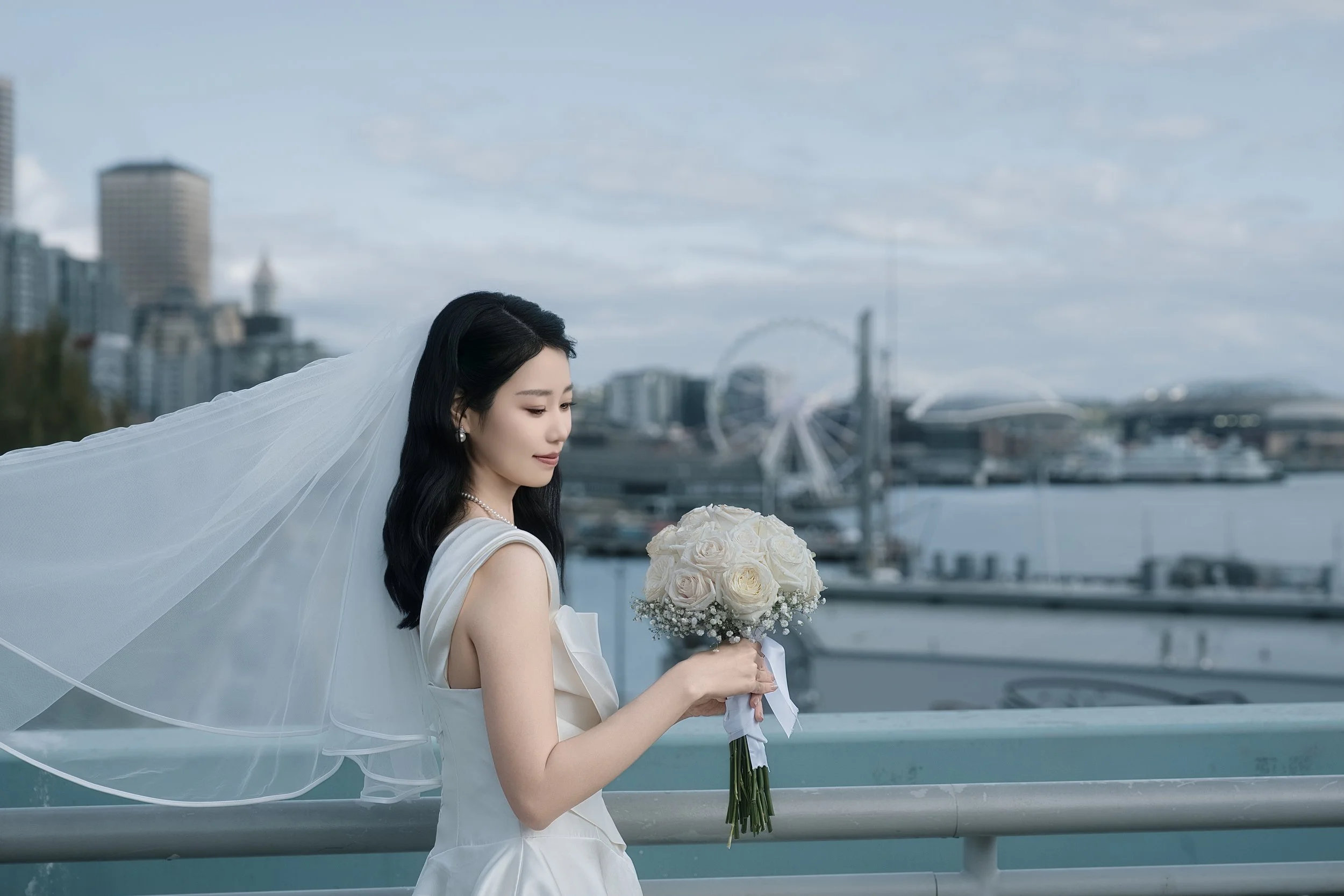 Bride in a white wedding dress and flowing veil holding a bouquet of white roses overlooking a waterfront city skyline