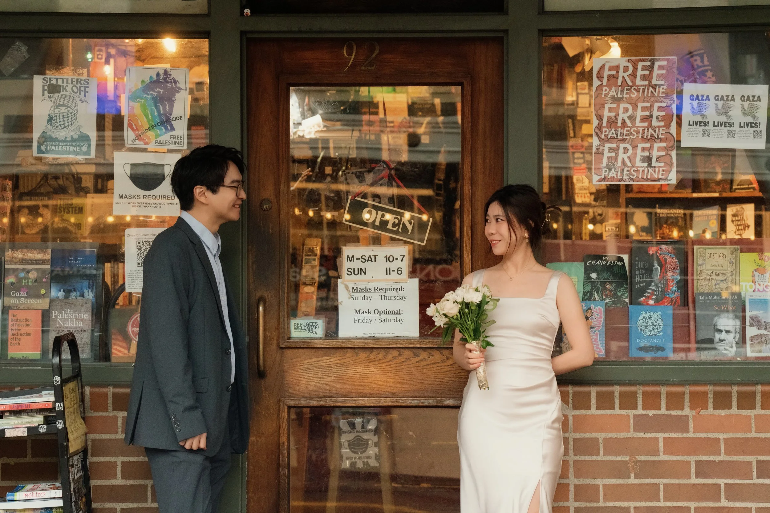 Couple posing outside a bookstore, with the woman holding a bouquet and Palestine solidarity posters displayed in the windows.