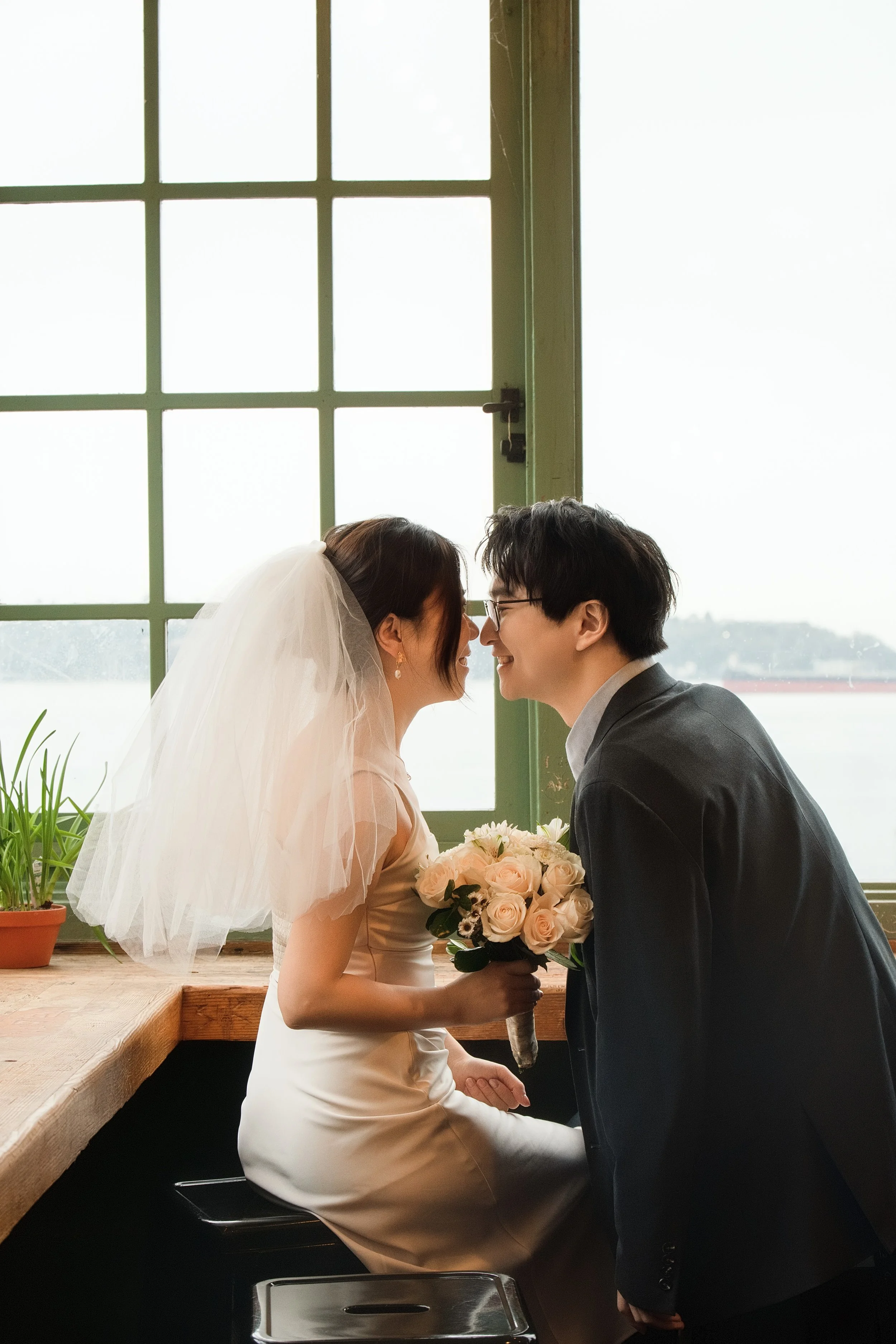 Bride and groom leaning in for a kiss beside a sunlit window, with the bride holding a bouquet of pale roses.