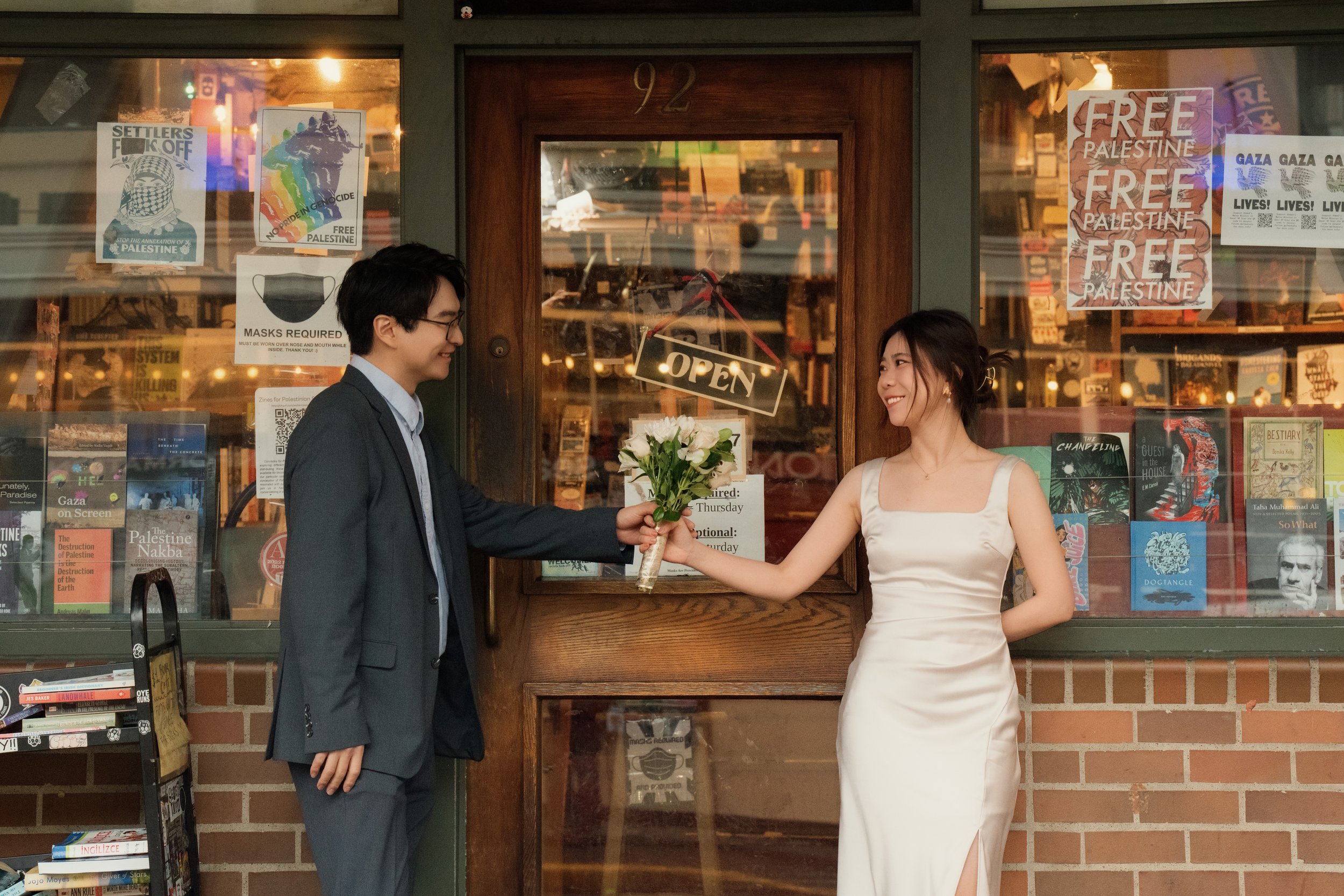 Man presenting a bouquet of white flowers to a smiling woman outside a bookstore decorated with Free Palestine posters