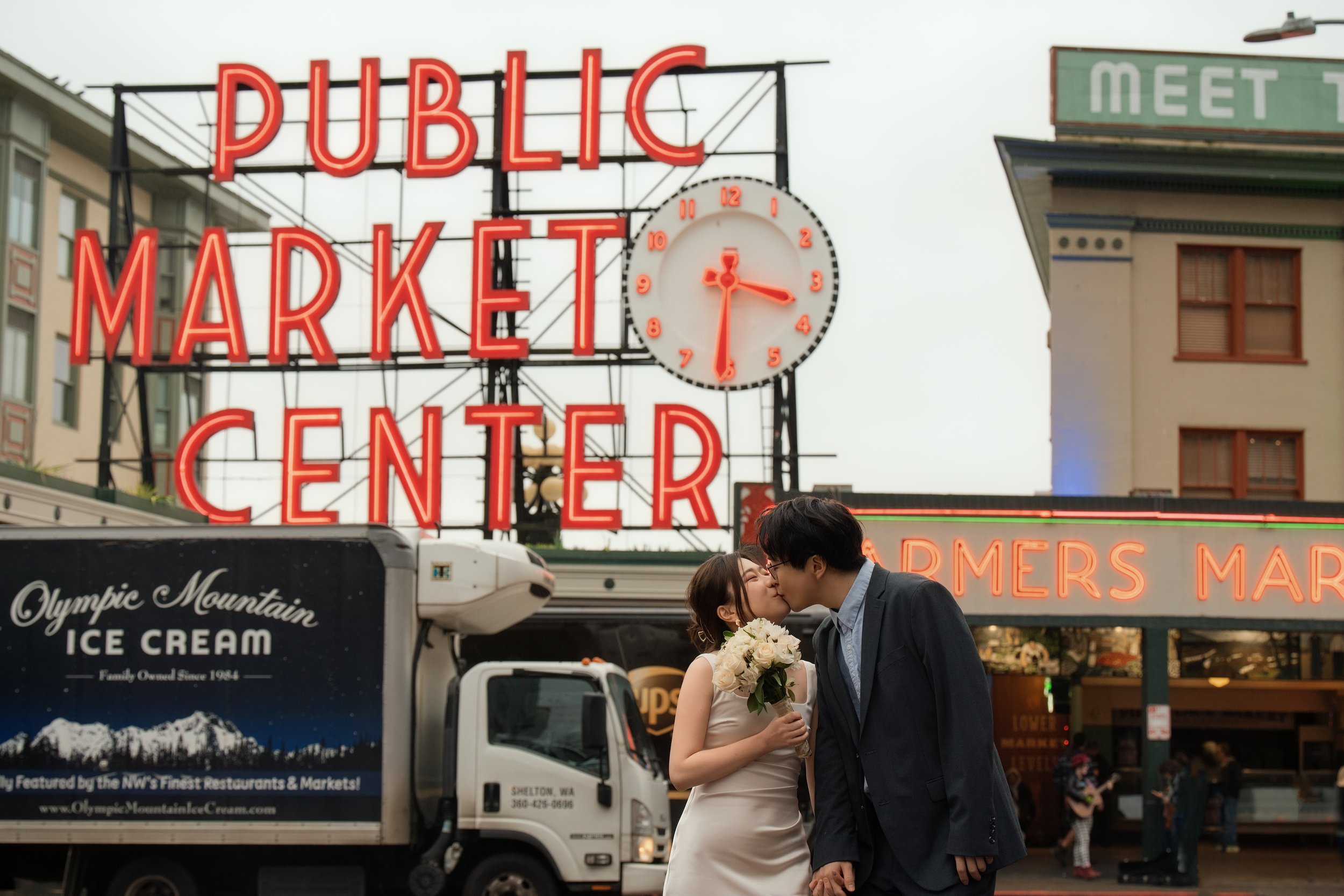 Newlywed couple kissing while holding hands and a bouquet beneath Pike Place Market's iconic neon sign in Seattle