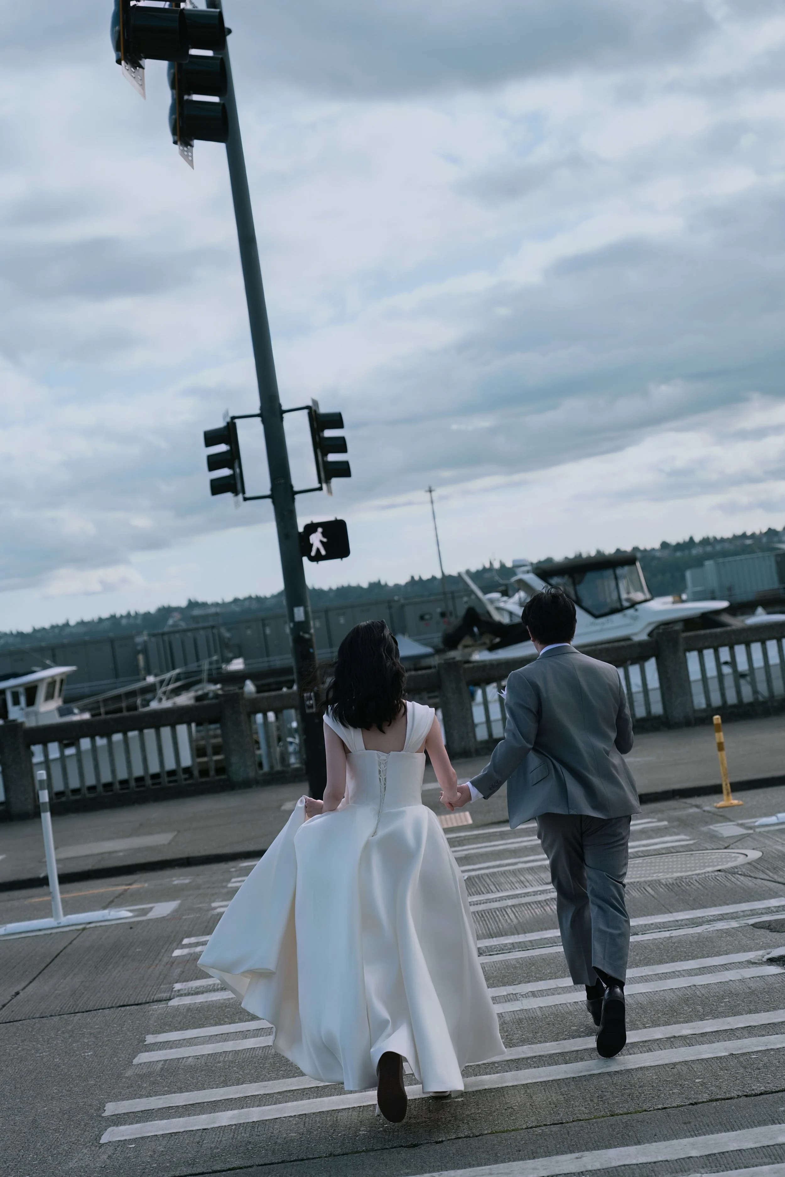 Bride and groom holding hands while crossing a waterfront street near boats and traffic lights