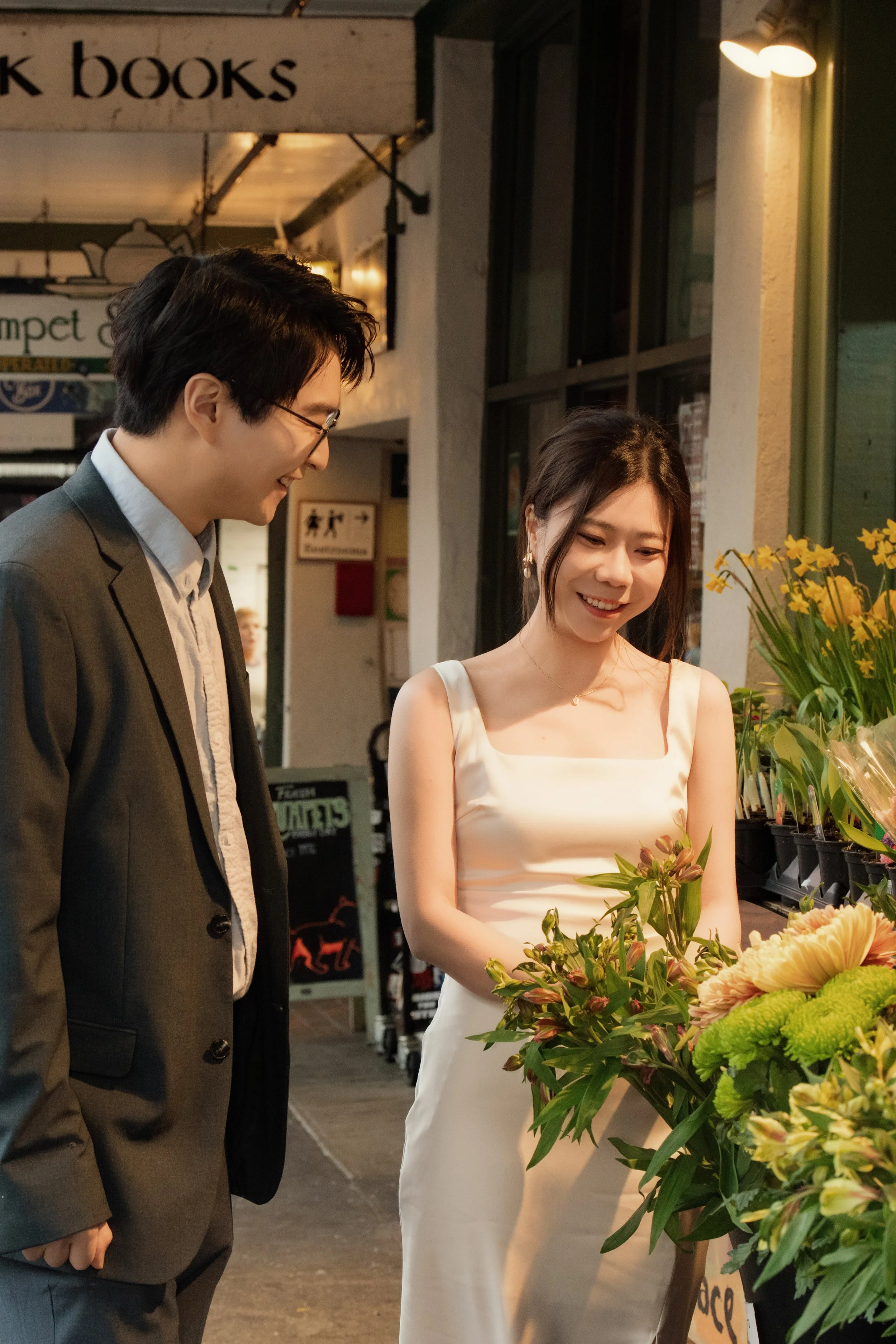 Smiling bride holding a colorful flower bouquet beside a groom in a suit outside a flower shop