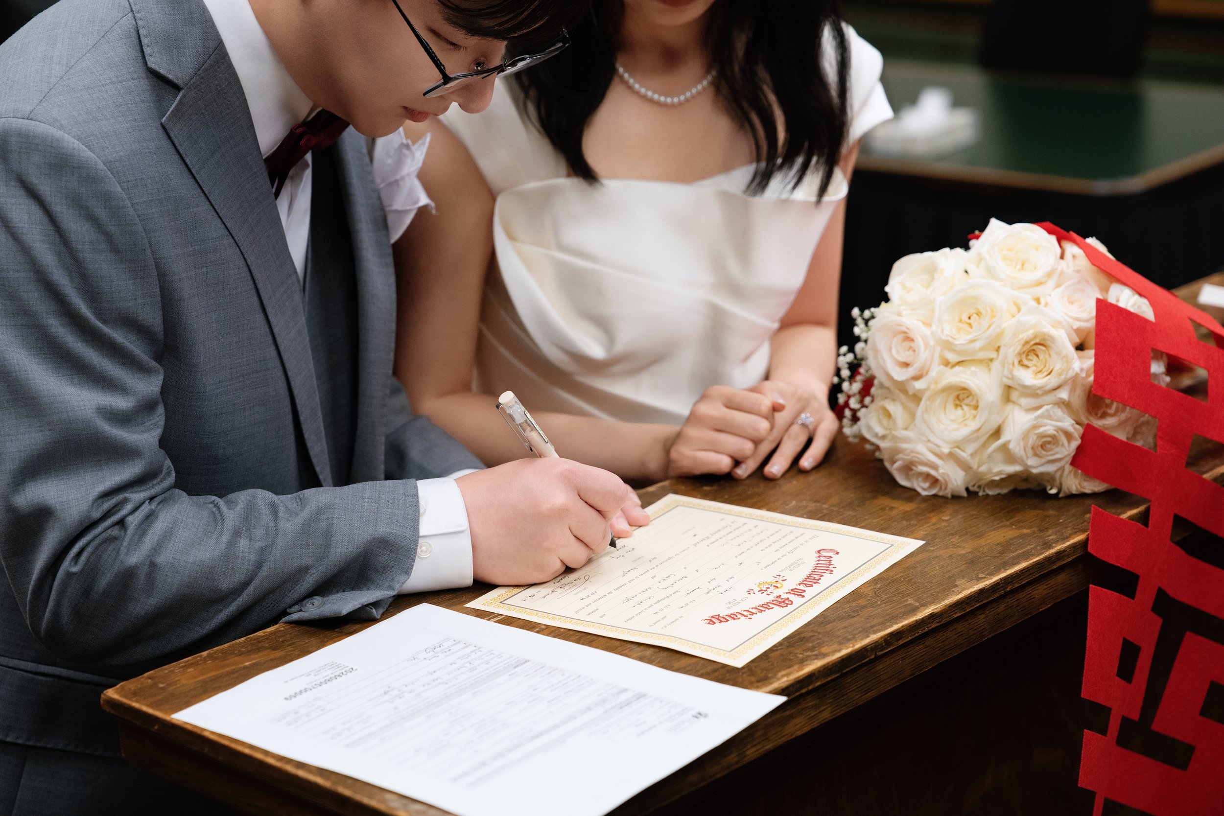Bride and groom signing a marriage certificate at a wedding ceremony beside a bouquet of white roses.