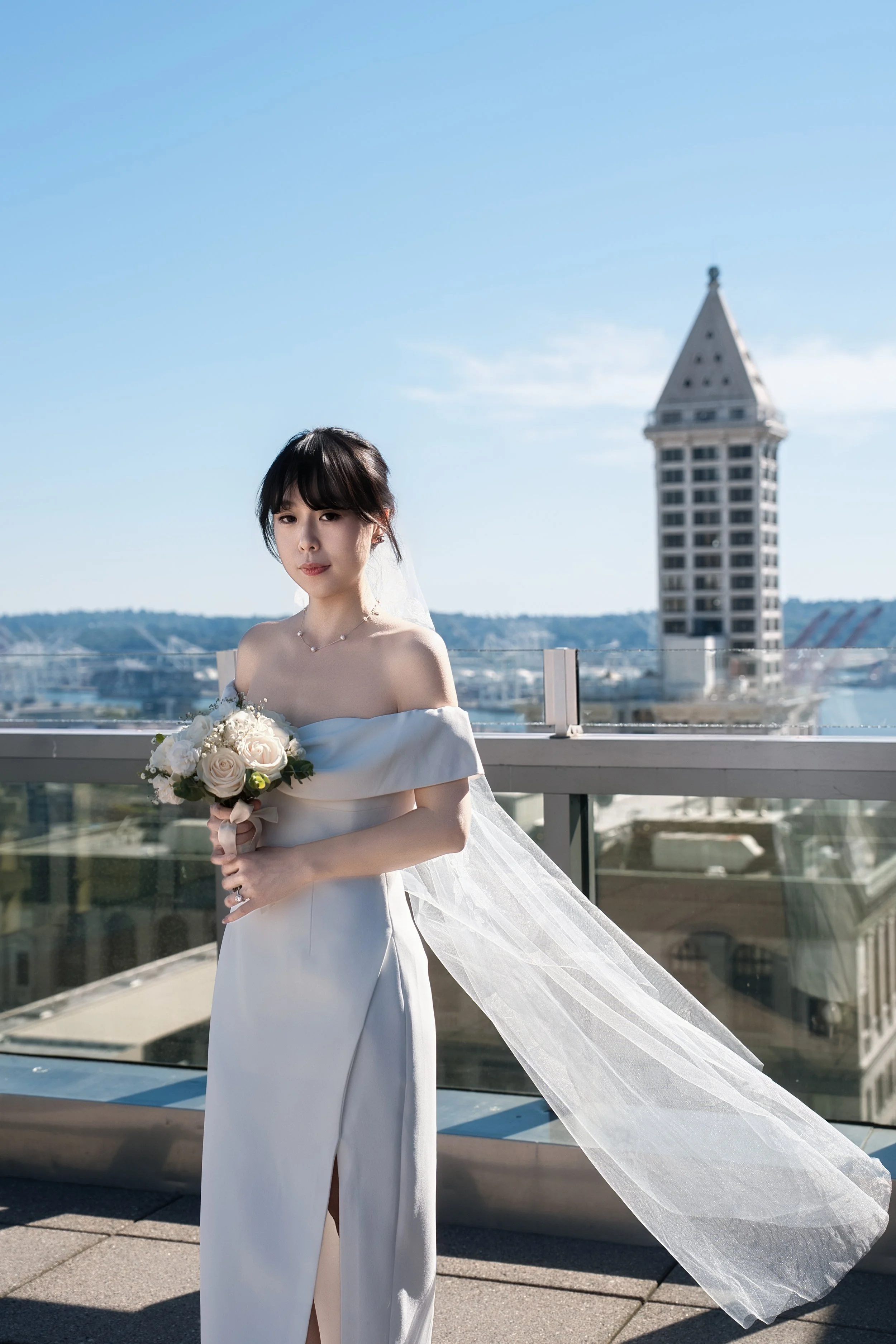 Bride in a white off-the-shoulder wedding dress holding a bouquet on a rooftop with a city skyline behind her