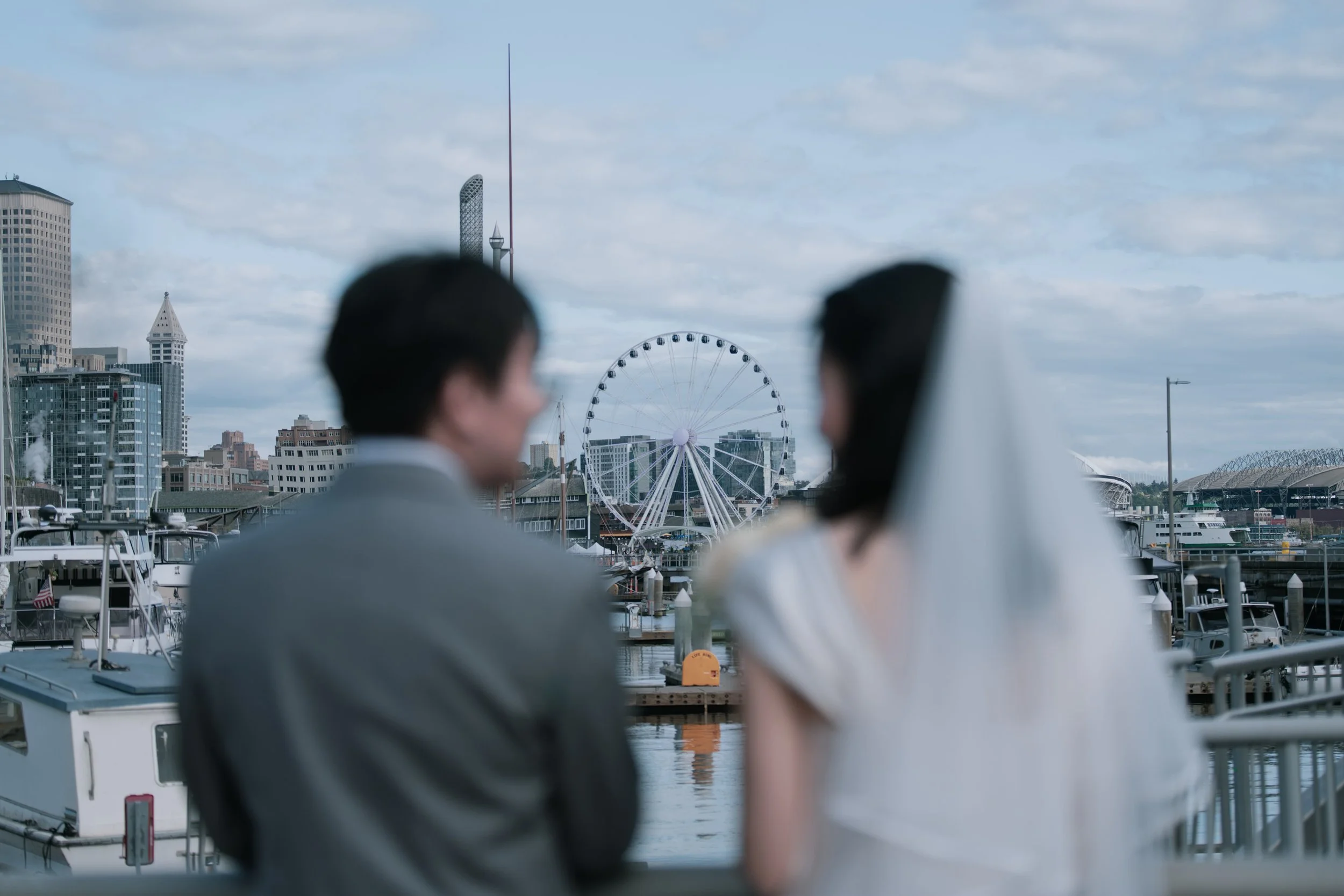 Blurred people overlooking Seattle's waterfront, harbor boats, the Seattle Great Wheel, and downtown skyline.