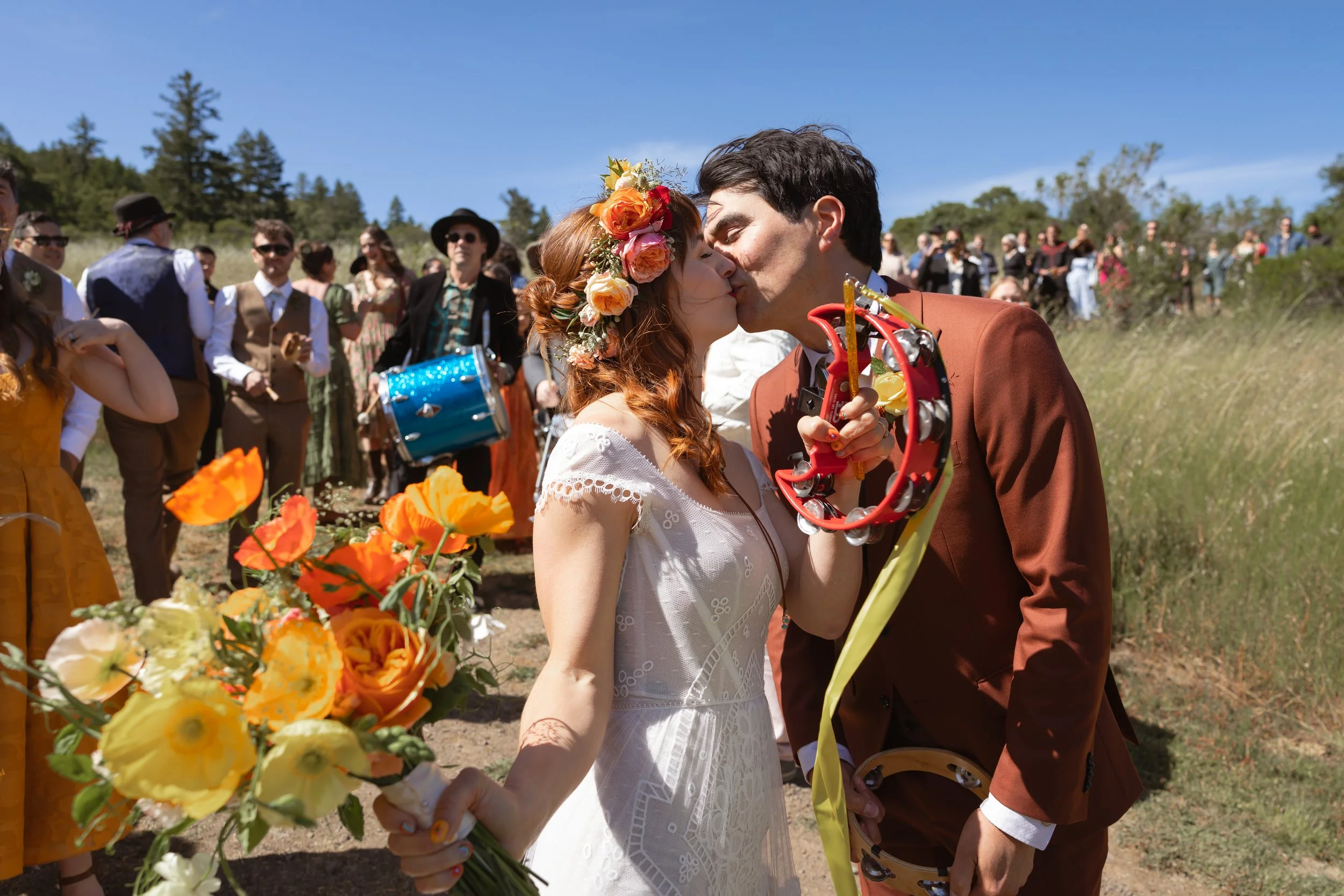 couple kissing while bride holds a bridal bouquet and tamborine in front of a procession outdoors