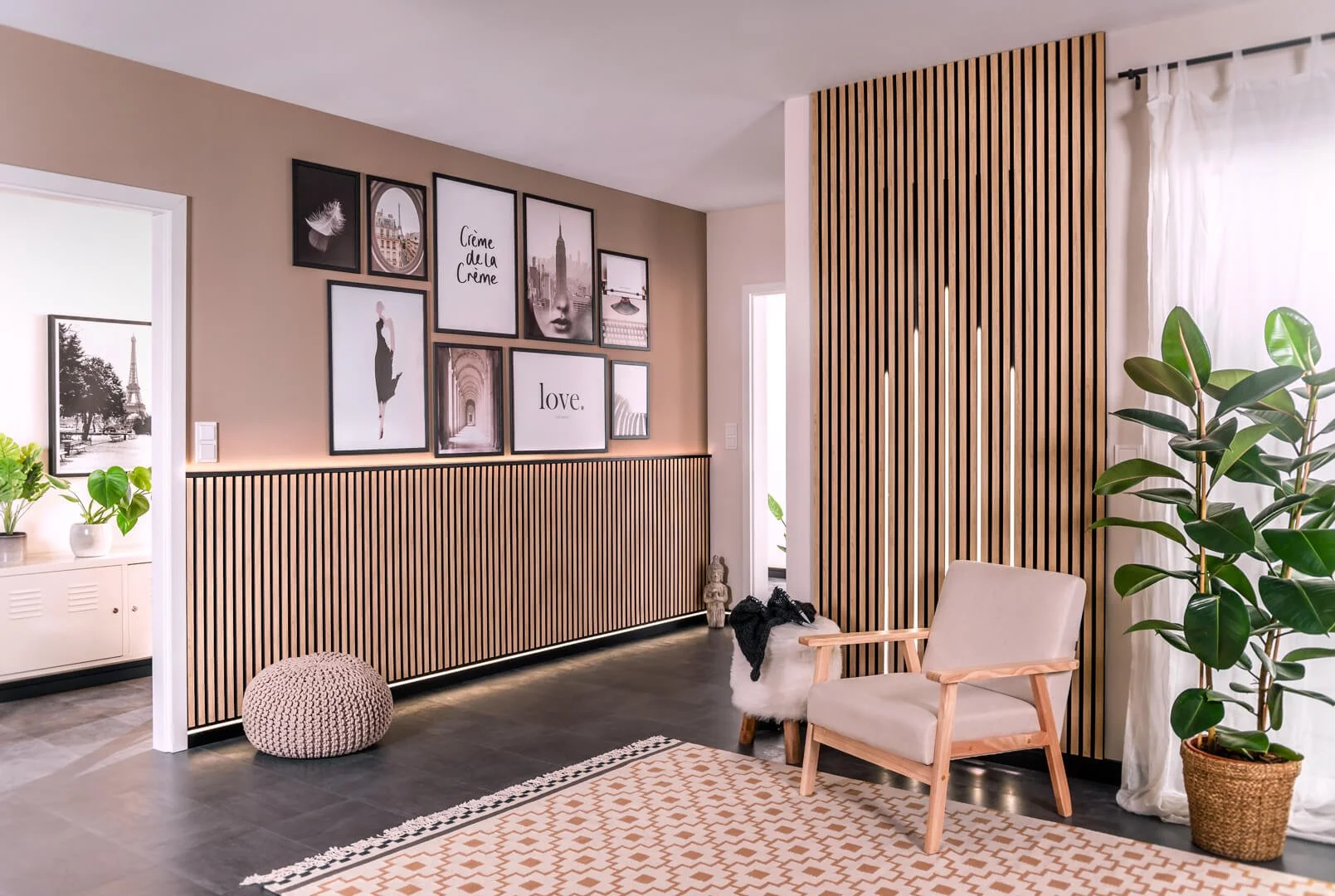 Modern living room with beige walls, framed artwork, and wooden slatted dividers. Contains a beige armchair, woven pouf, potted plant, and patterned rug.