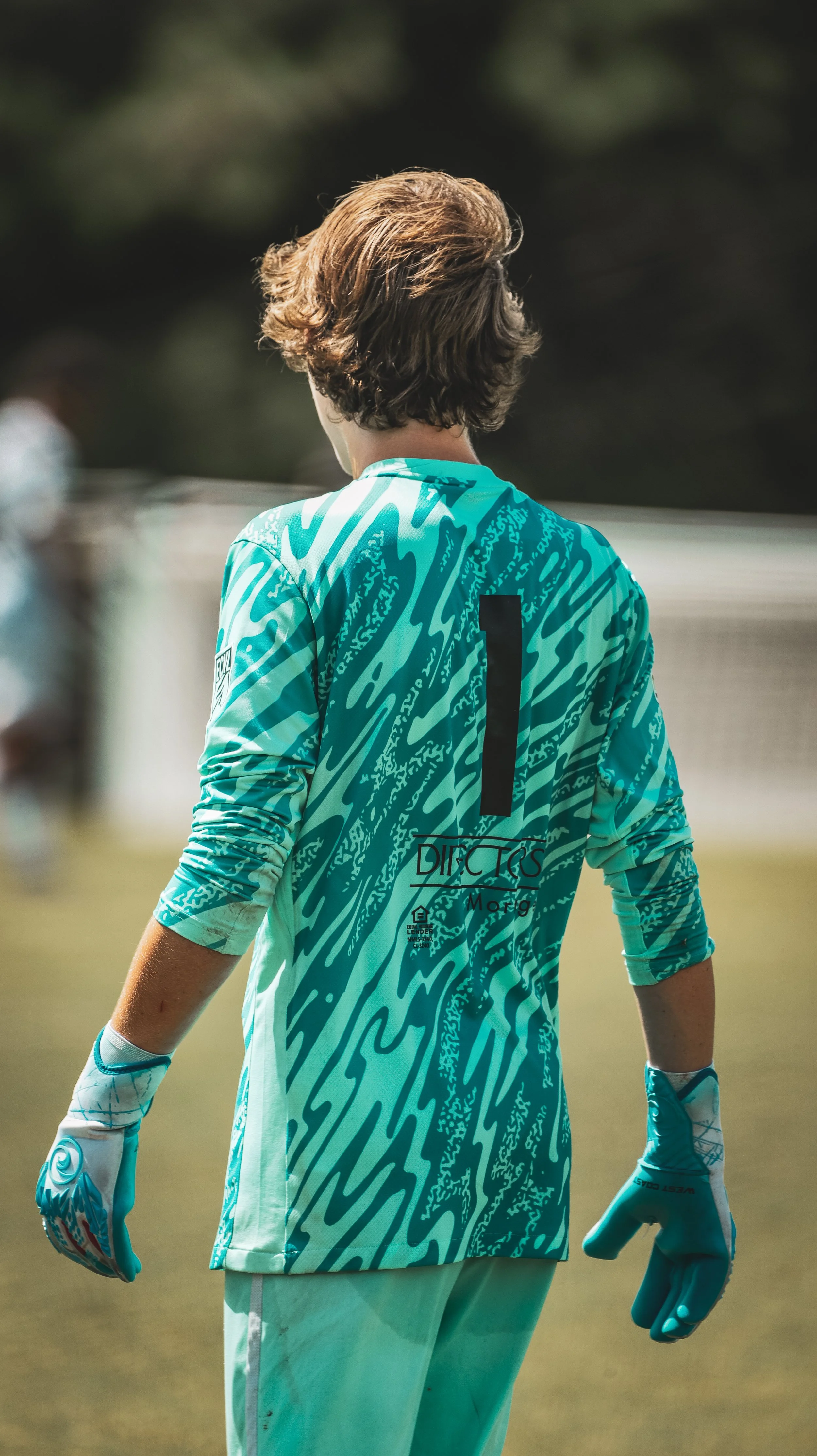 Back view of a female soccer goalkeeper wearing a turquoise and black patterned jersey with the number 1 on back, and matching gloves, standing on the field.