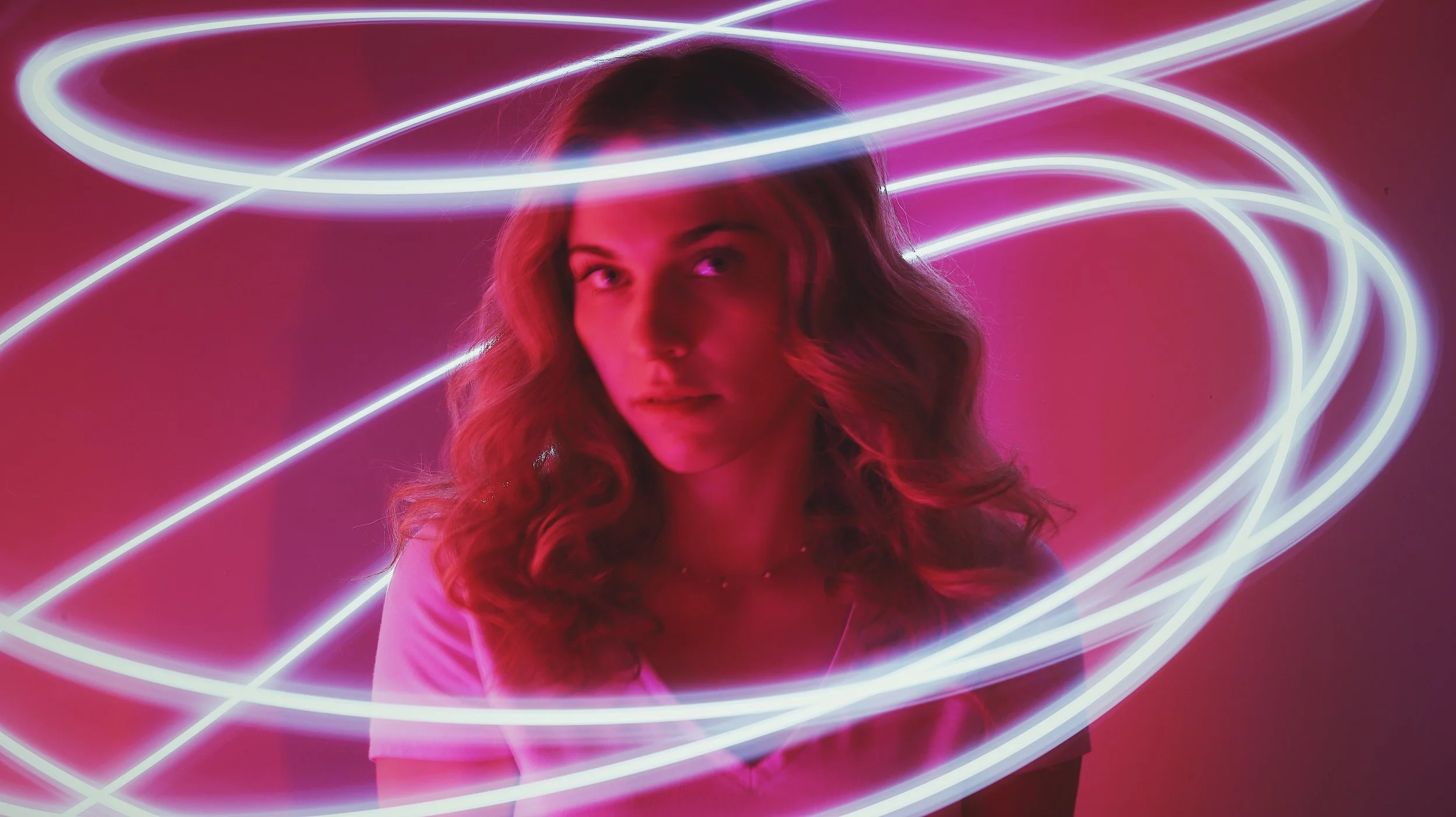 A woman with wavy hair is surrounded by pink and white neon light streaks in a dark room, with a pink background.