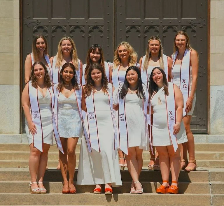 A group of eleven young women in white dresses standing on steps in front of a large metal door, wearing white sashes with red and purple Greek letters and the words "Class of 2024".