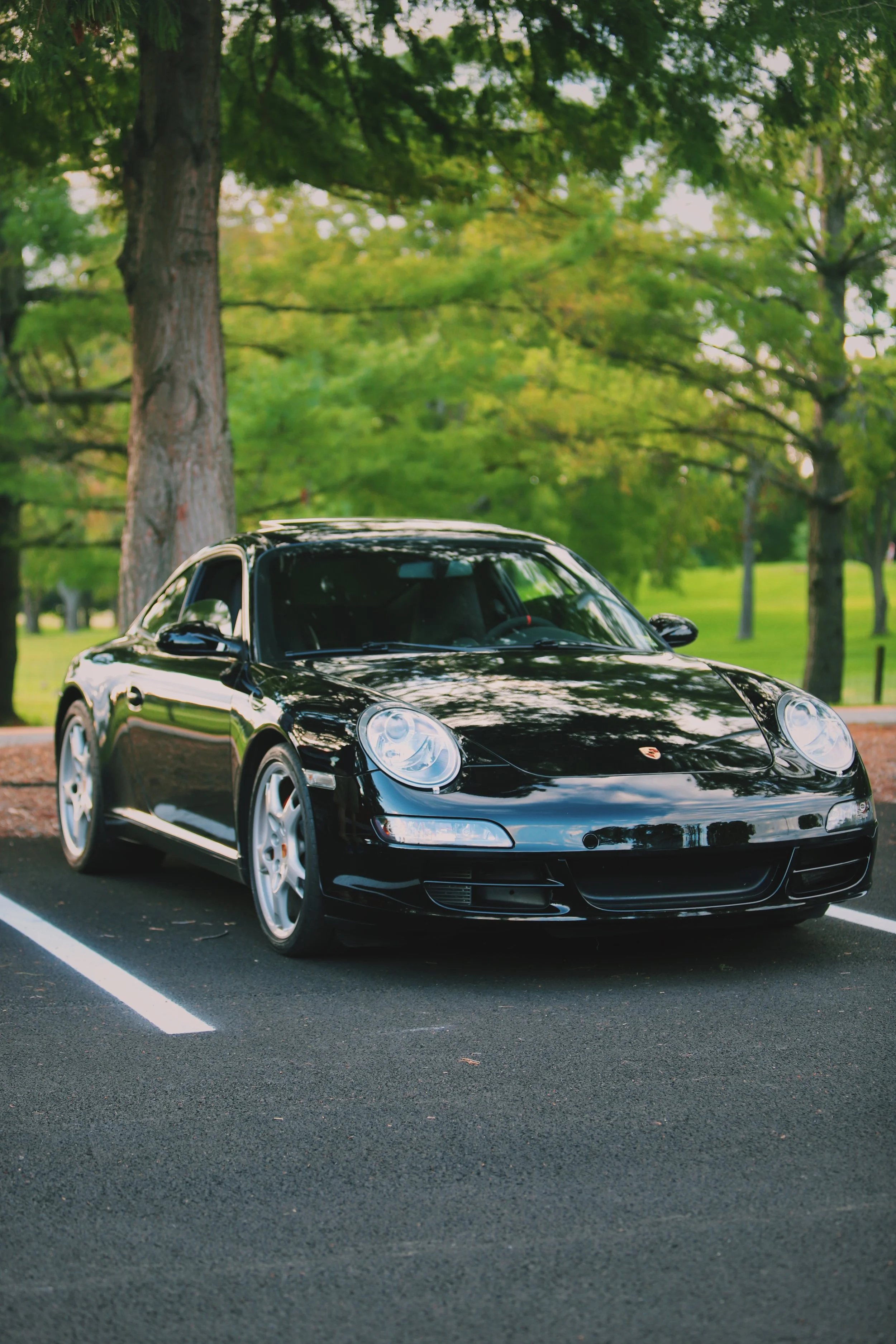 Black Porsche sports car parked in a parking lot with trees and greenery in the background.