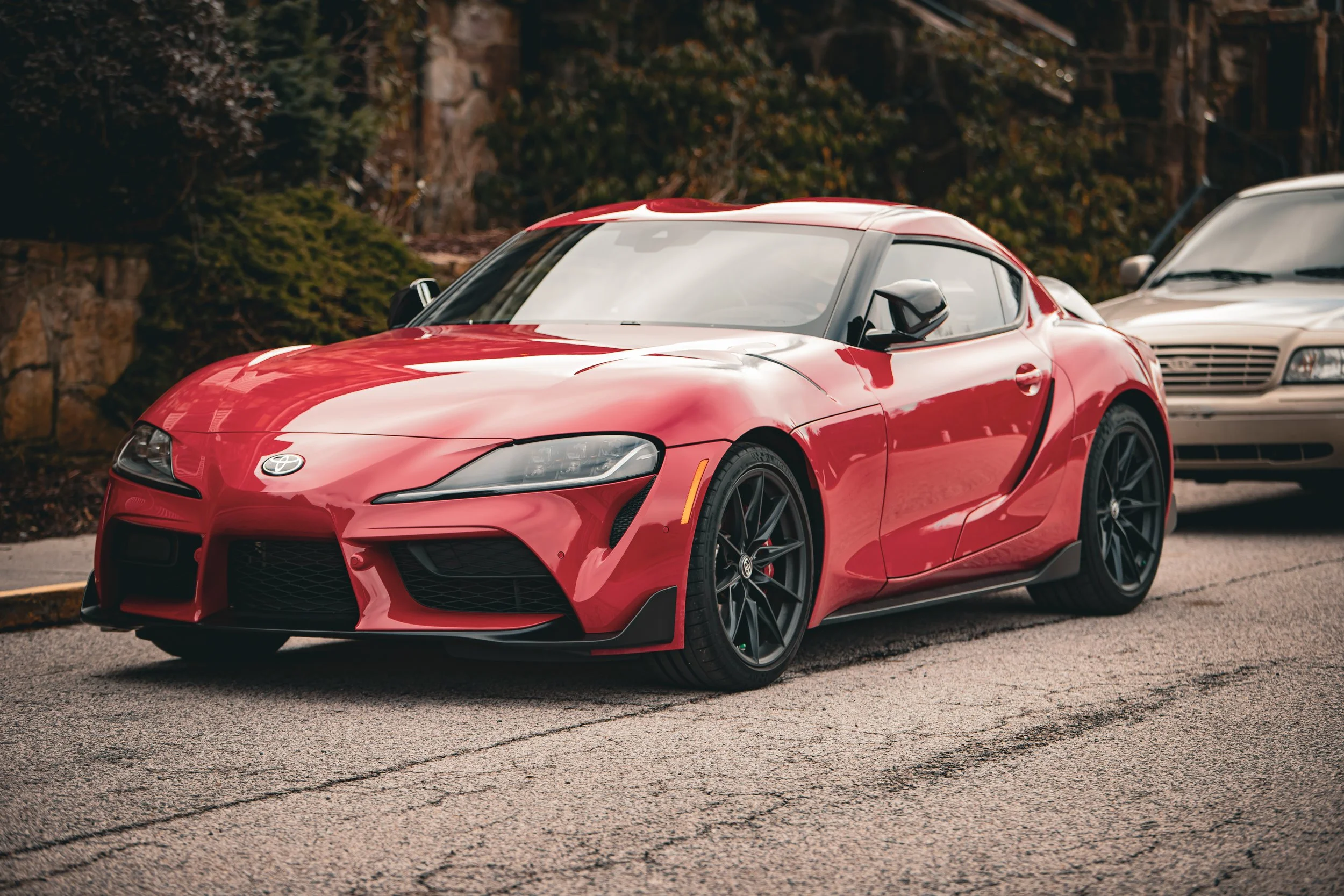 A red sports car parked on the street with a second beige car behind it, trees, rocks, and bushes in the background.