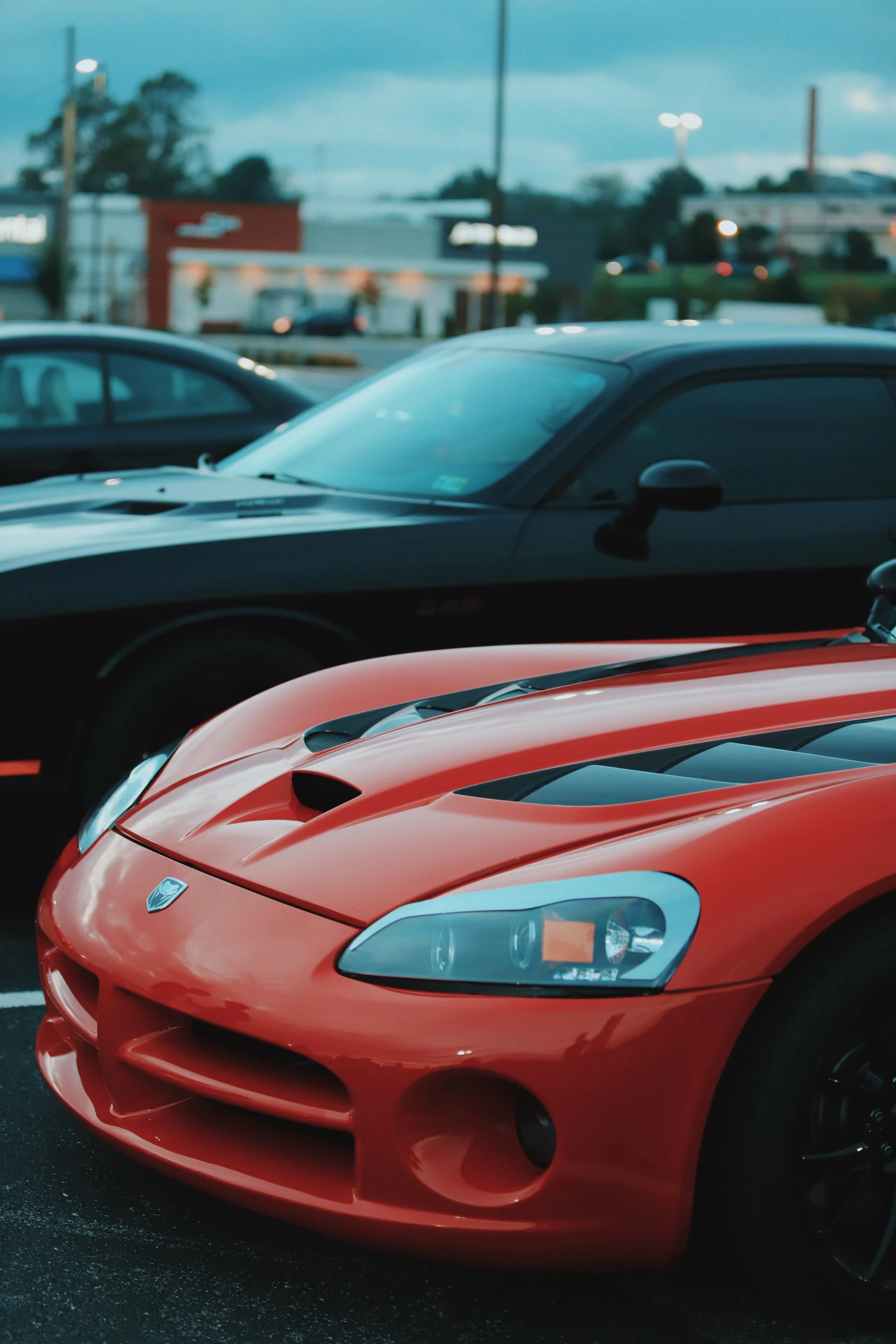 Close-up of a red Dodge Viper sports car parked in a lot, with other cars and a shopping center visible in the background during dusk.