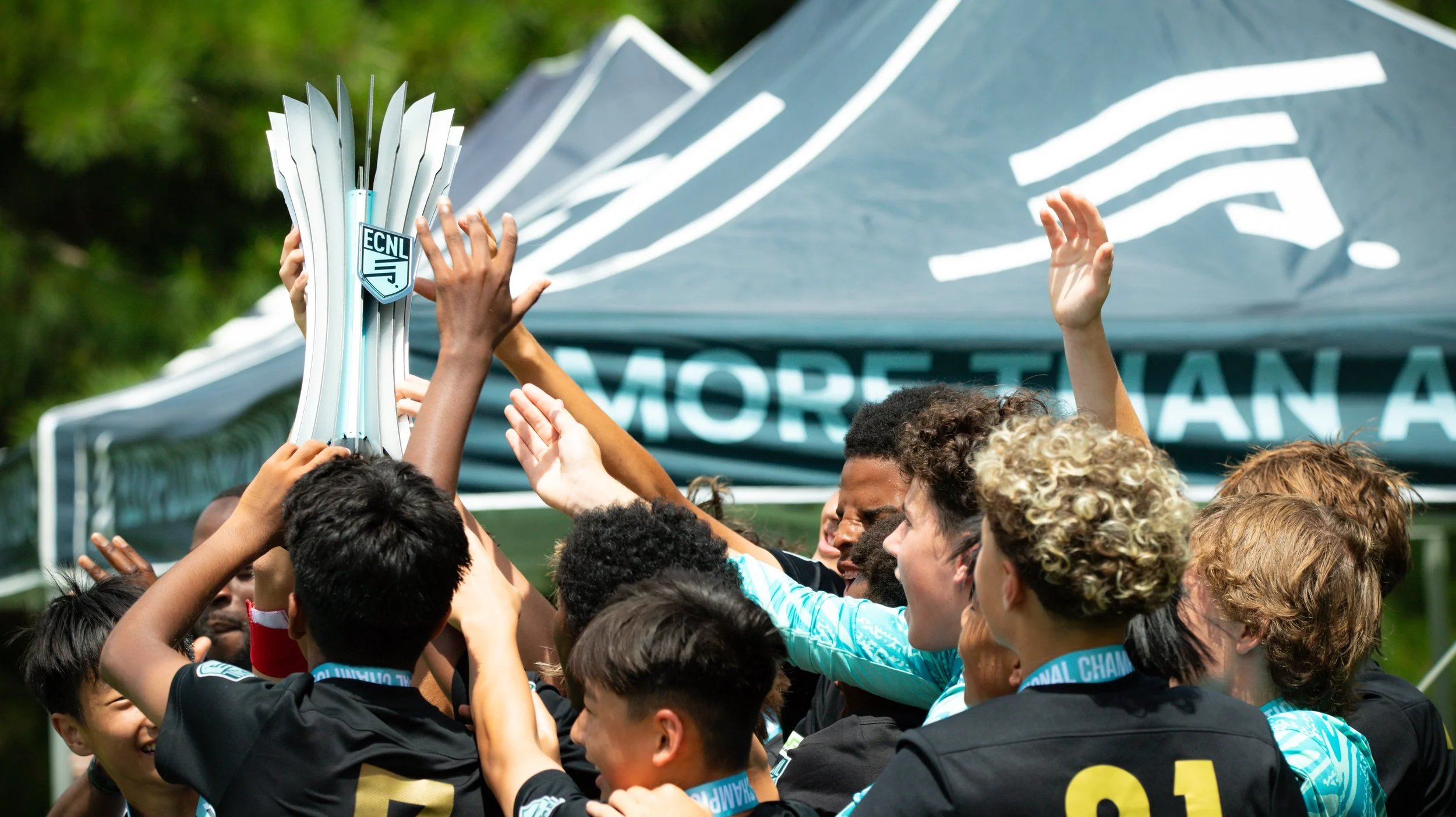 A diverse group of young soccer players celebrating and lifting a trophy in front of a tent with the words 'MORE THAN' visible.
