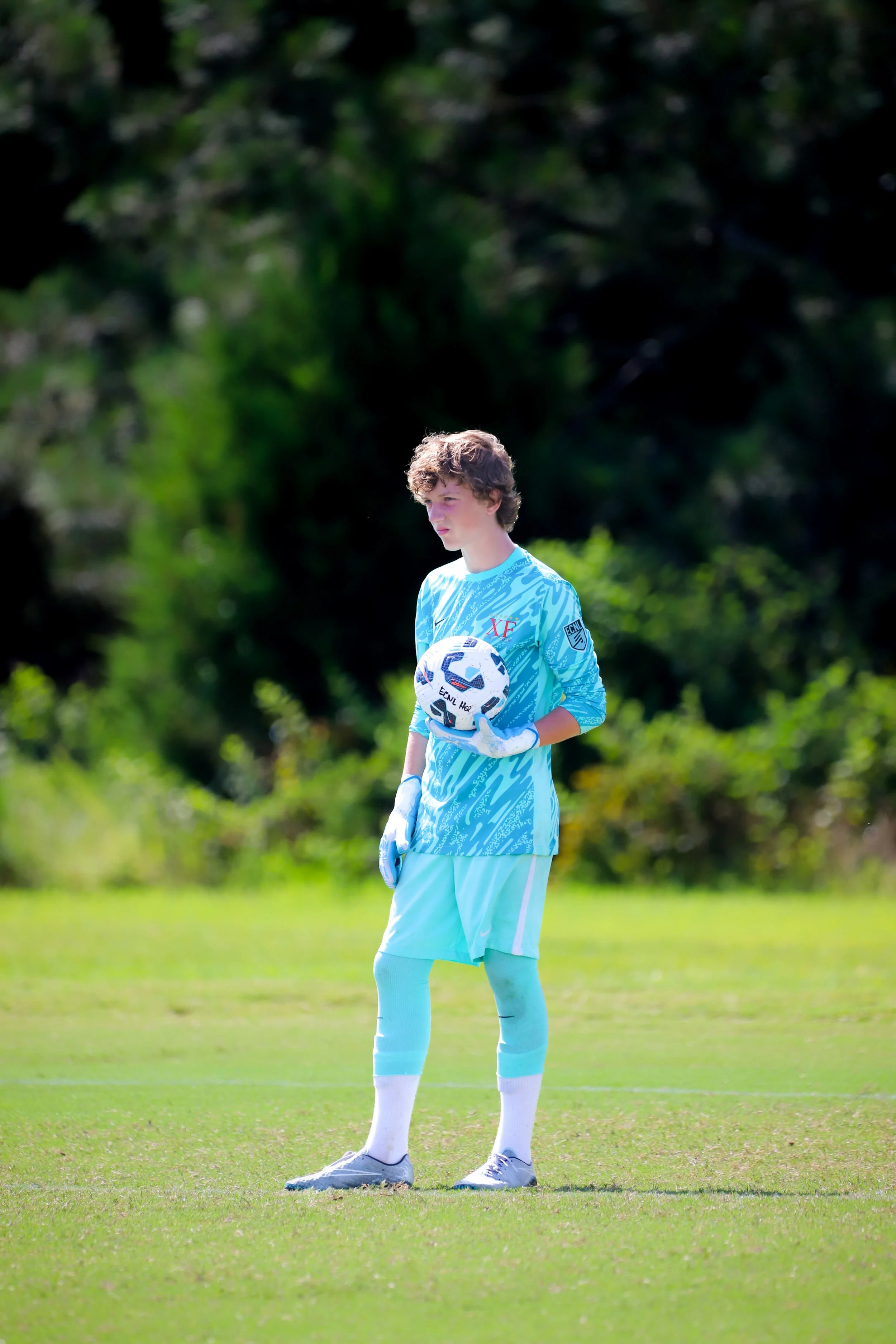 A young male soccer goalkeeper on the field, holding a soccer ball, dressed in a blue goalkeeper uniform with gloves, standing on a grassy field with trees in the background.