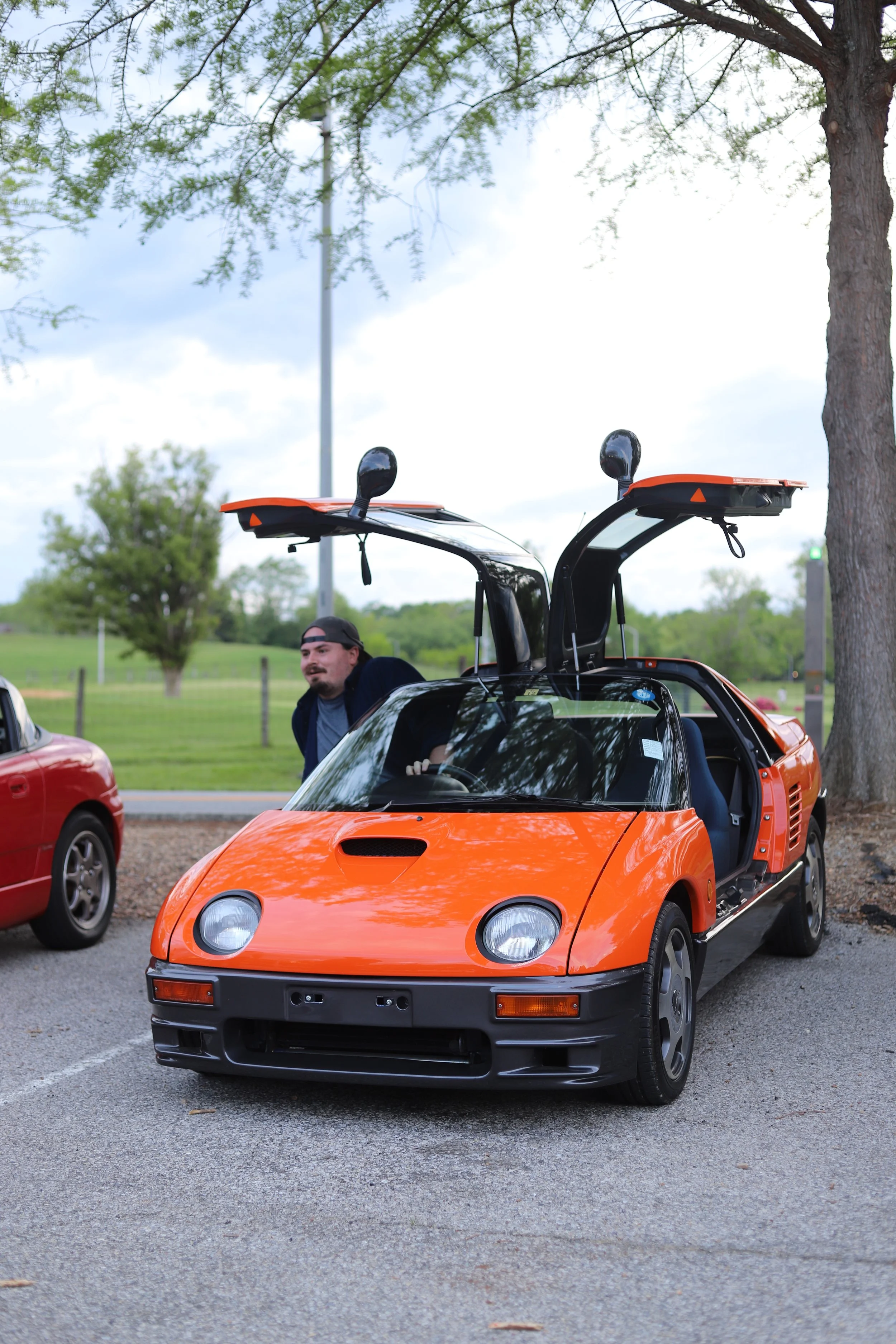 A man standing next to a bright orange vintage sports car with gullwing doors open, parked in a lot with a grassy field, trees, and a cloudy sky in the background.